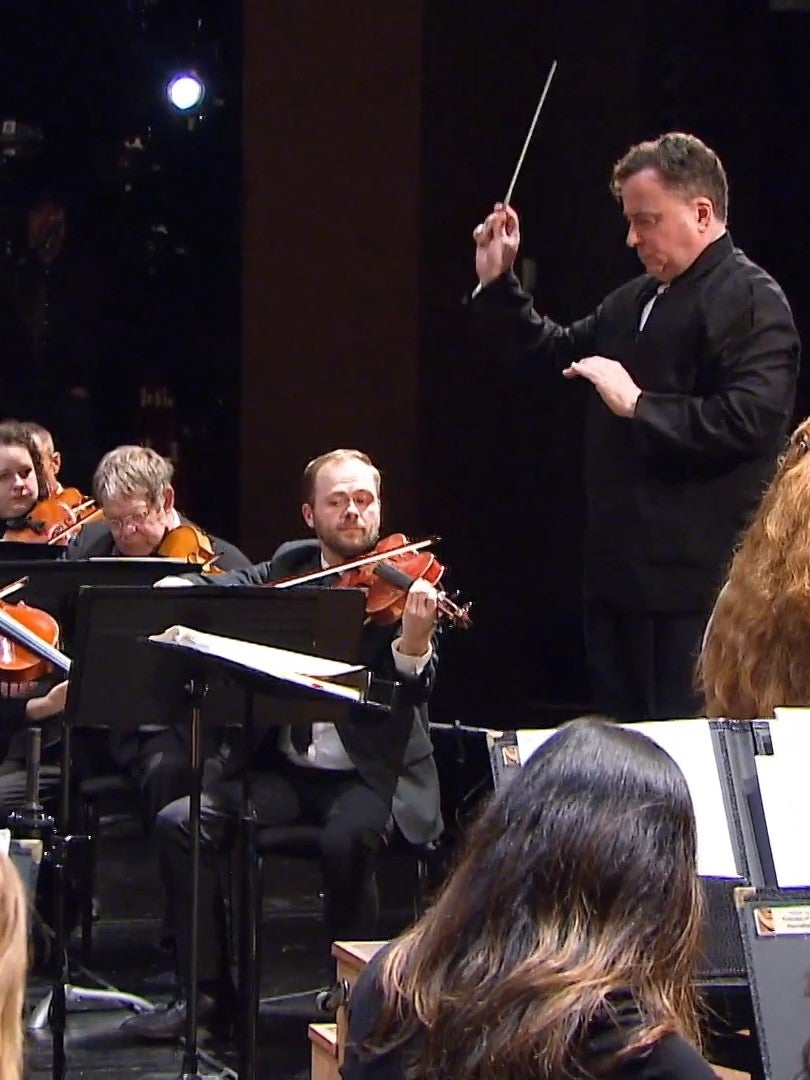 A conductor leads an orchestra as violinists play their instruments during a live performance on stage.