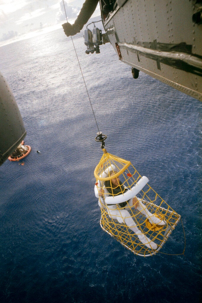 A person in a white suit is being lifted in a yellow rescue basket from the ocean by a hoist attached to a helicopter. An orange life raft floats nearby.