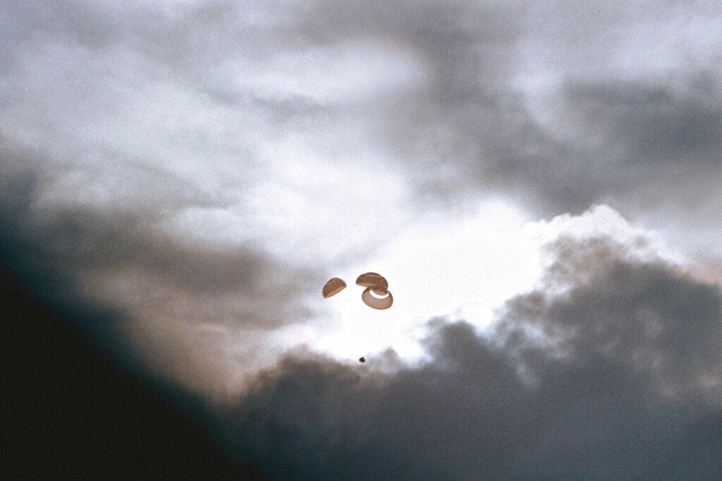 Three parachutes descend through a cloudy sky with a small object suspended below them, partially silhouetted against sunlight.