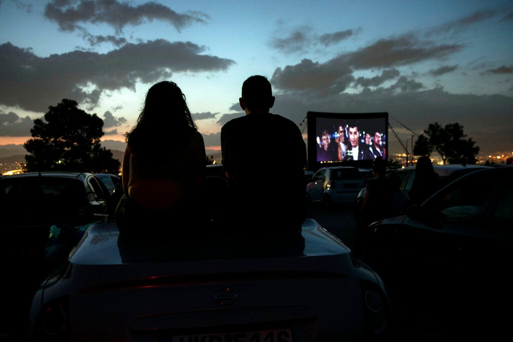 Two people sit on the roof of a car at a drive-in theater, watching a movie on an outdoor screen at dusk with parked cars around them.