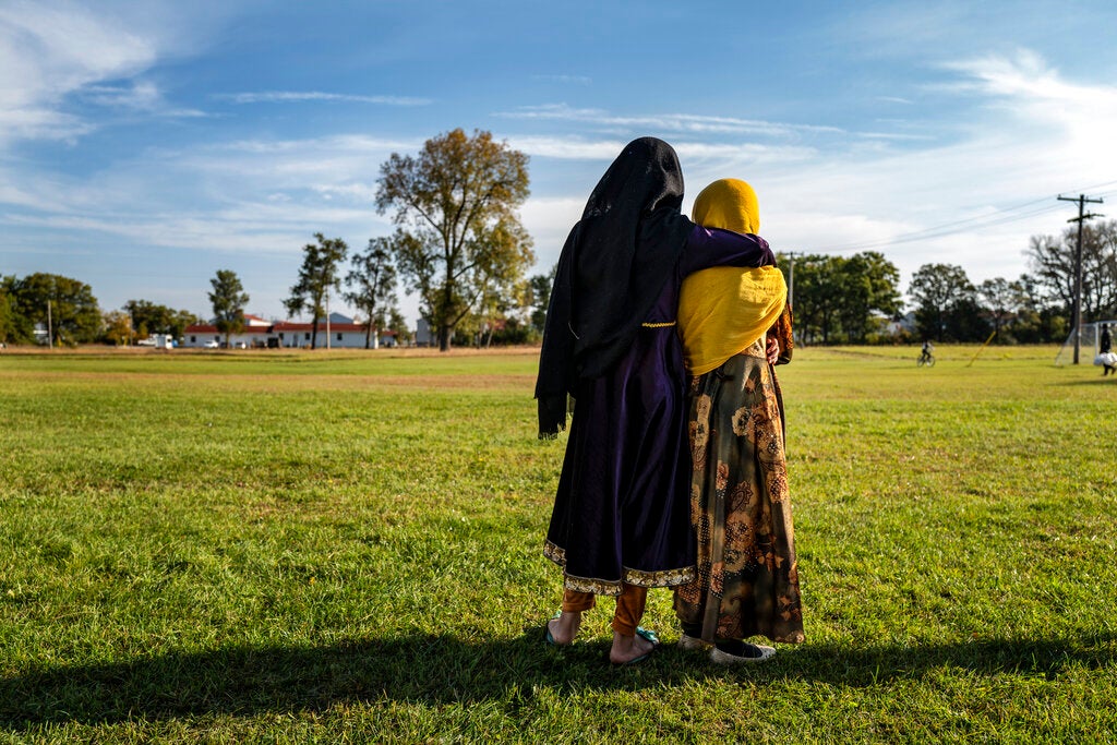 Two people wearing patterned dresses and headscarves stand together on a grassy field with their backs to the camera, one with an arm around the other. Trees and buildings are in the background.