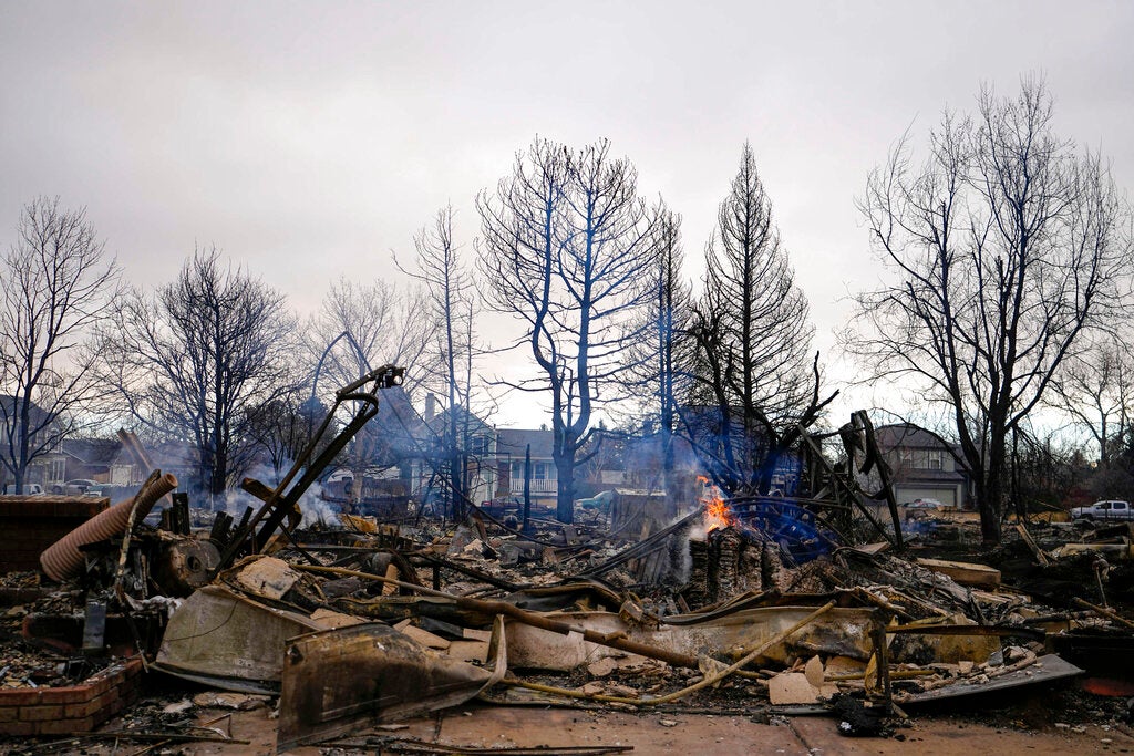 Charred debris and rubble remain after a fire, with smoldering flames and leafless trees in the background under a cloudy sky.