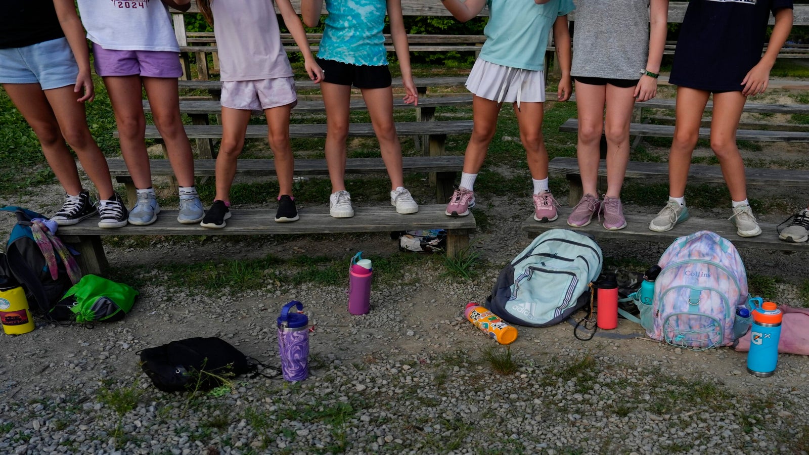 A group of children stand and sit on wooden bleachers with backpacks and water bottles on the gravel ground in front of them. Their faces are out of frame.