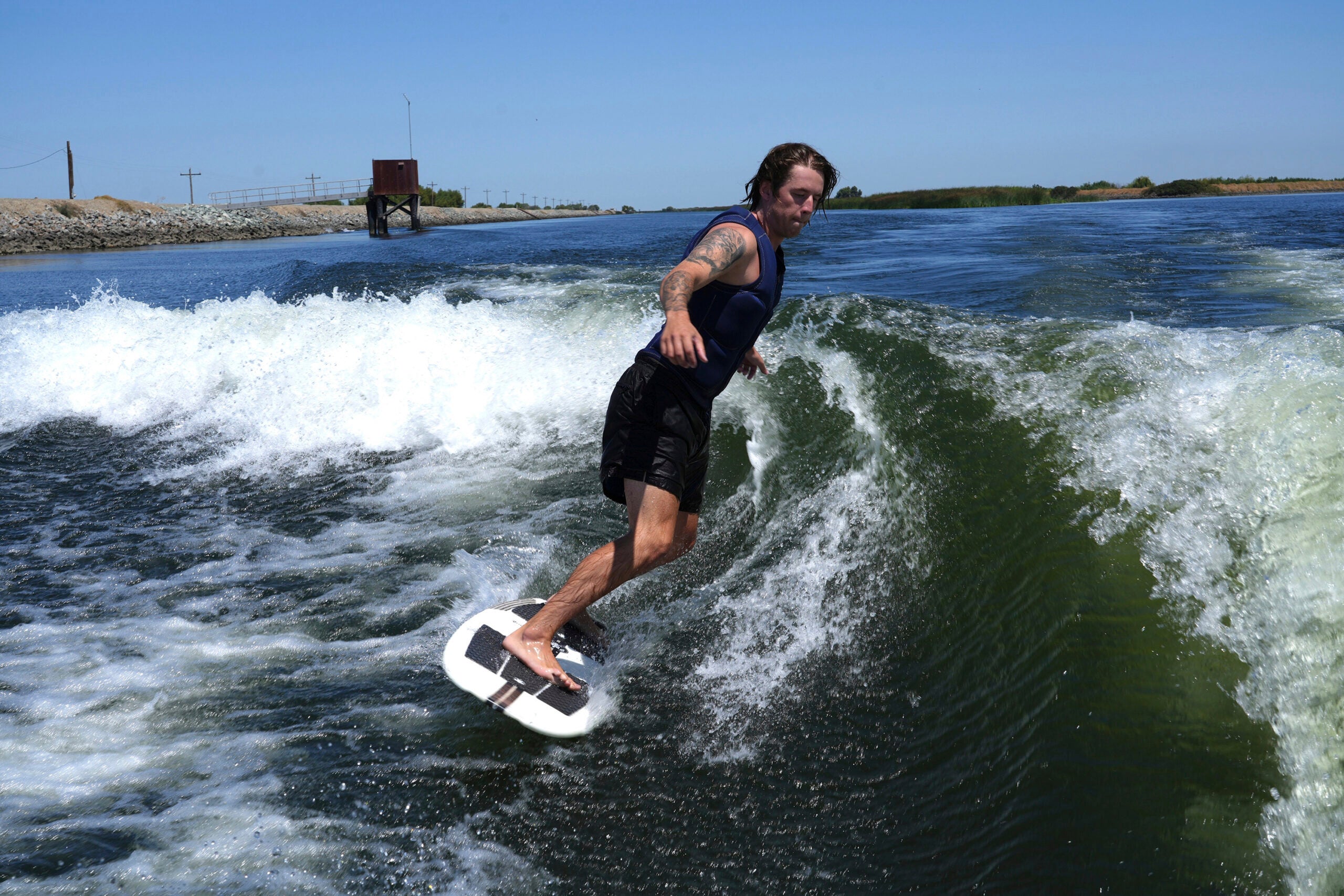 A person with tattoos on their left arm rides a wakeboard on a river, balancing on a wave under a clear blue sky.
