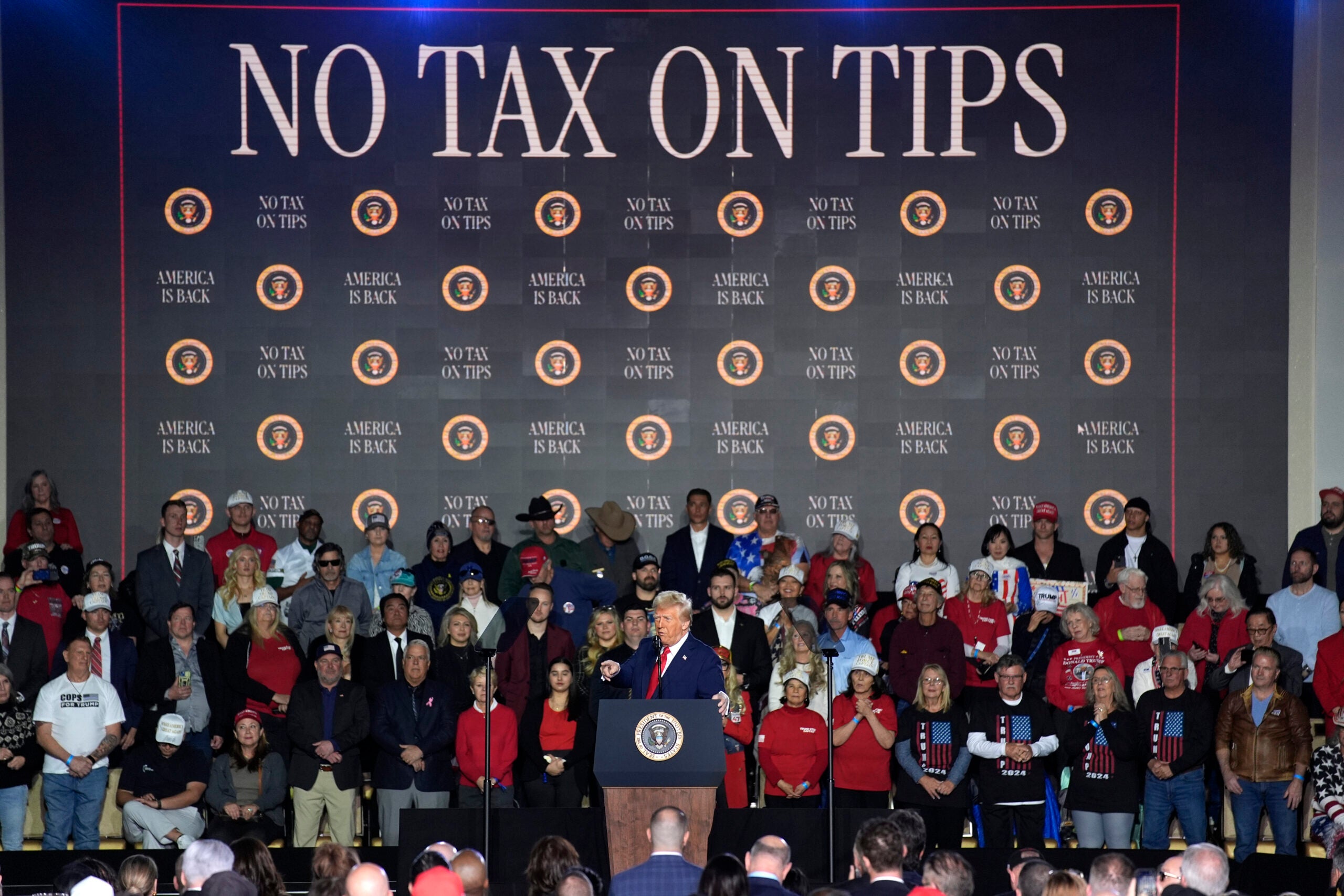 A crowd stands on stage behind a podium with a presidential seal; a large backdrop reads NO TAX ON TIPS repeatedly in bold letters.