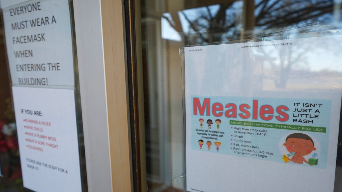 A printed sign about measles symptoms is taped to a glass door next to another notice requiring face masks before entering the building.