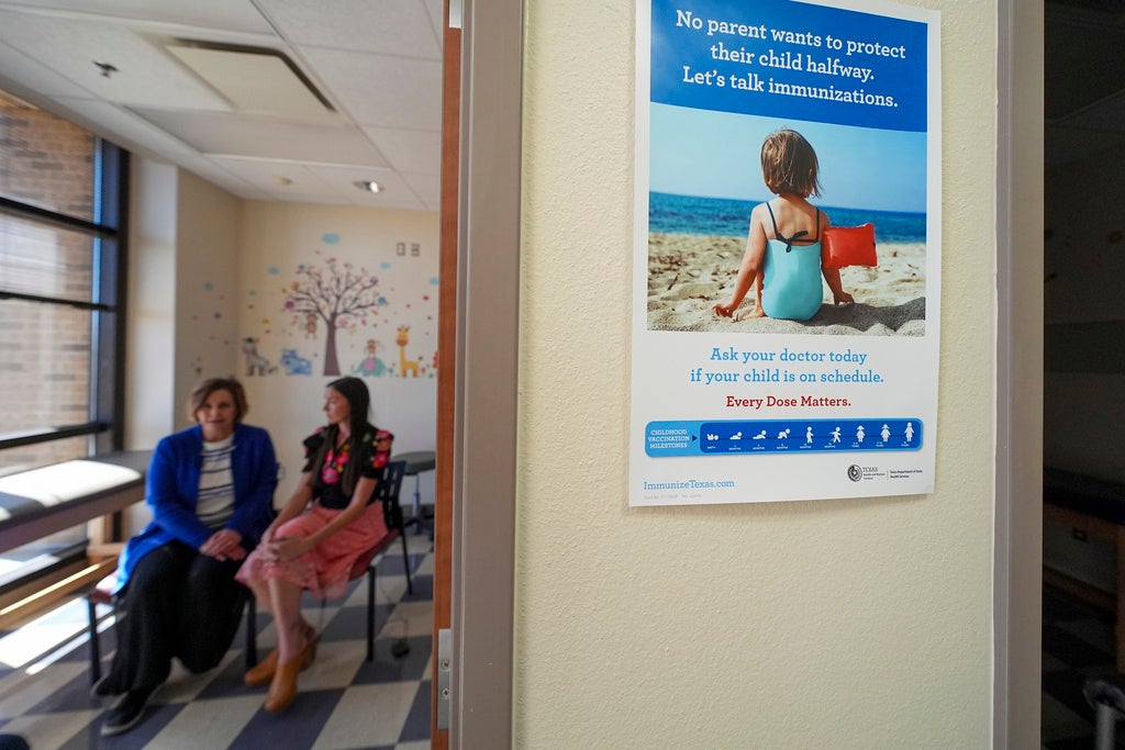 A poster about child immunizations is displayed on a wall in a clinic, with two women sitting and talking in the background by a window.