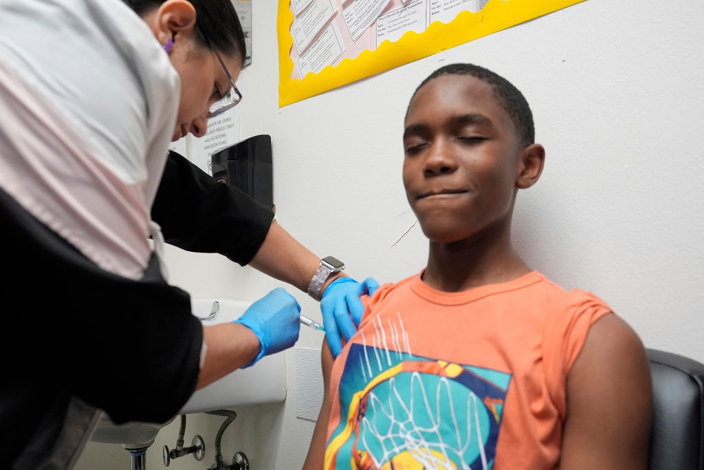A healthcare worker administers a vaccine to a boy wearing an orange shirt who sits with his eyes closed and mouth slightly pursed.