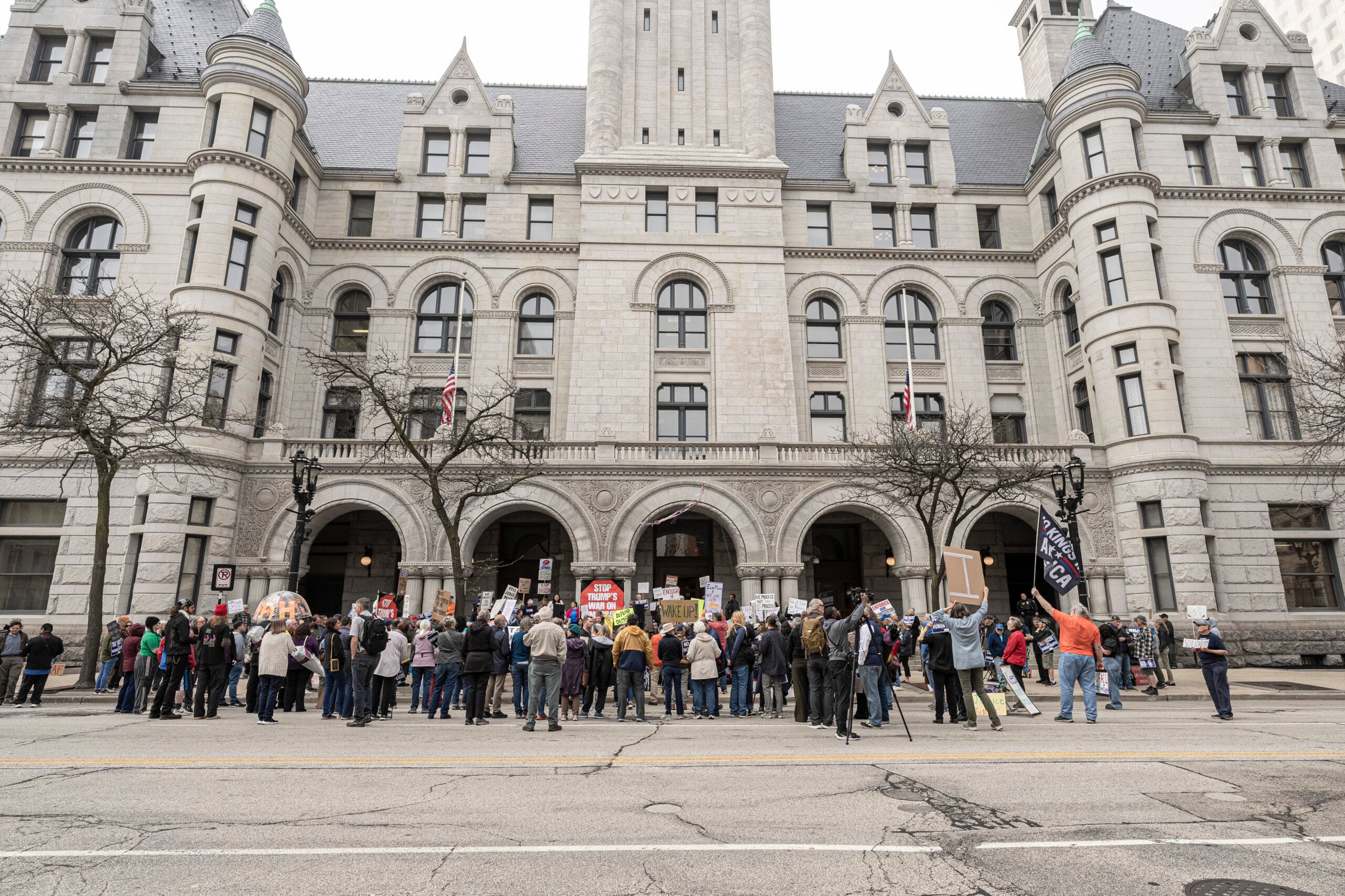 A group of people holding signs gather in protest outside a large historic stone building with arched entrances and towers.