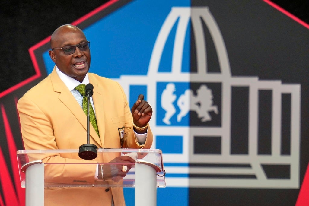 A man in a beige jacket speaks at a podium in front of a graphic backdrop featuring a stylized building and football player silhouettes.