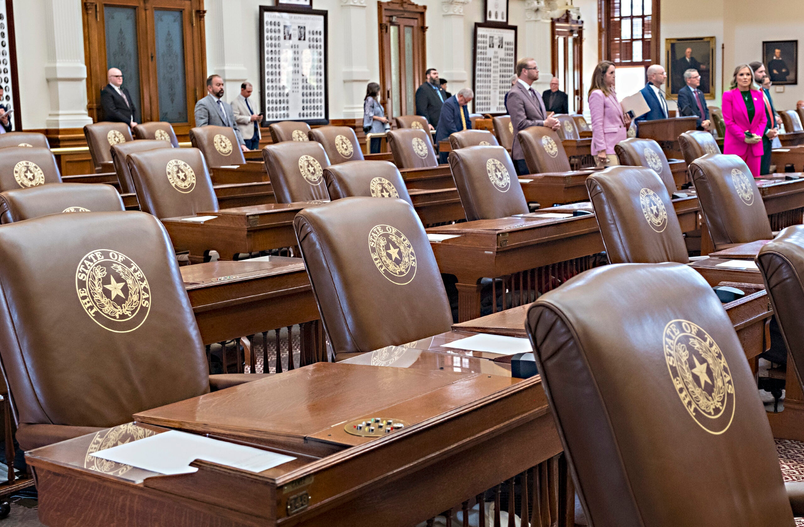 Rows of empty leather chairs with the Texas state seal in a legislative chamber, with several people standing and talking in the background.