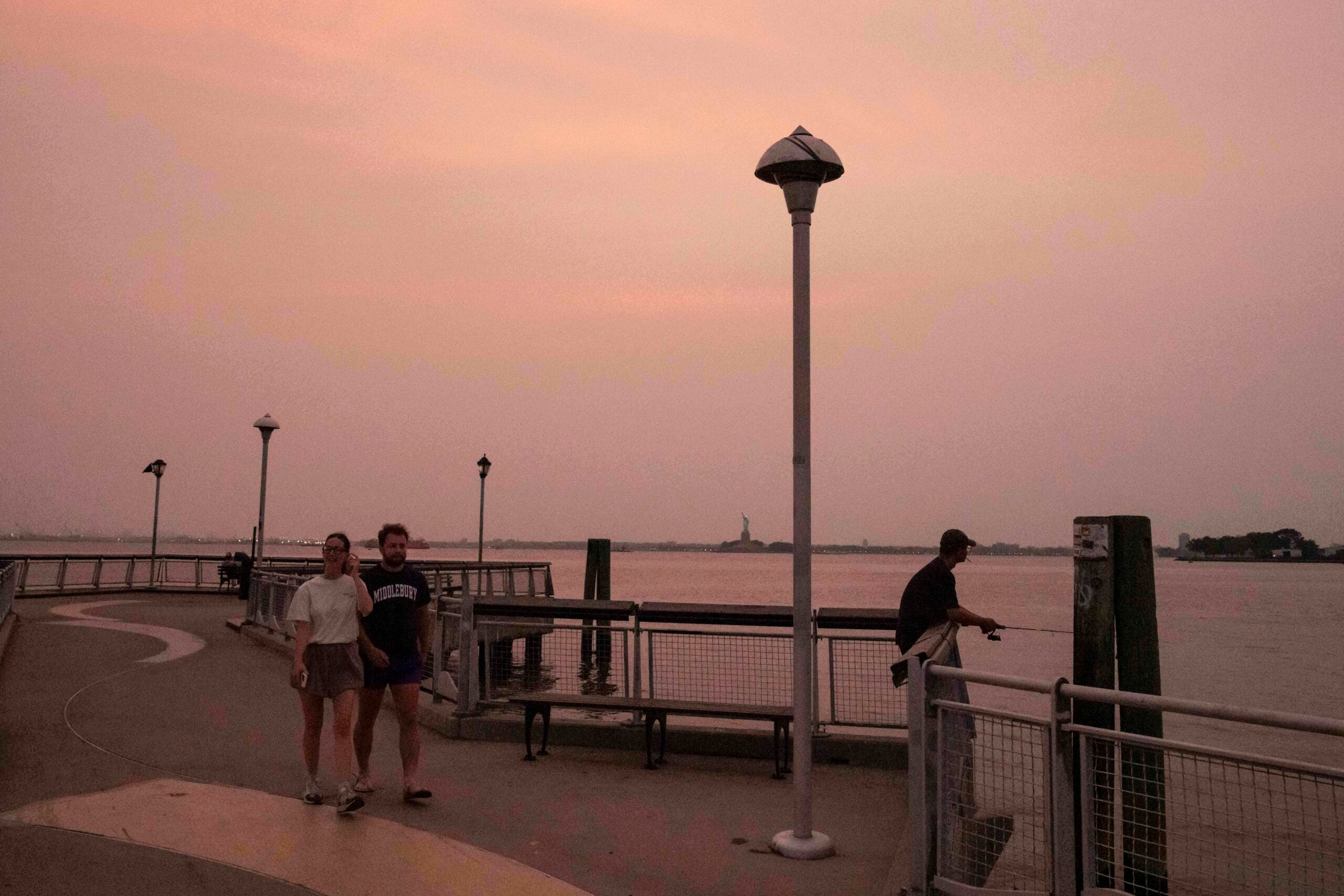 Two people walk along a riverside path at sunset, while another person fishes by the railing under a pink-orange sky.