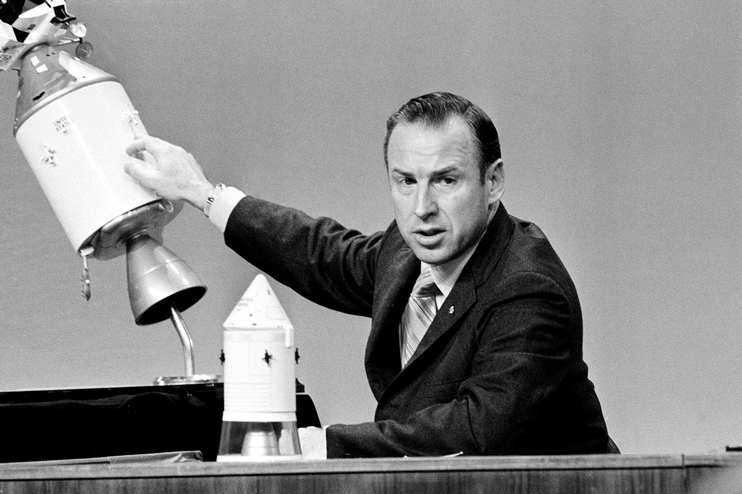 A man in a suit demonstrates with a model of a spacecraft, holding one section apart from another on a table.