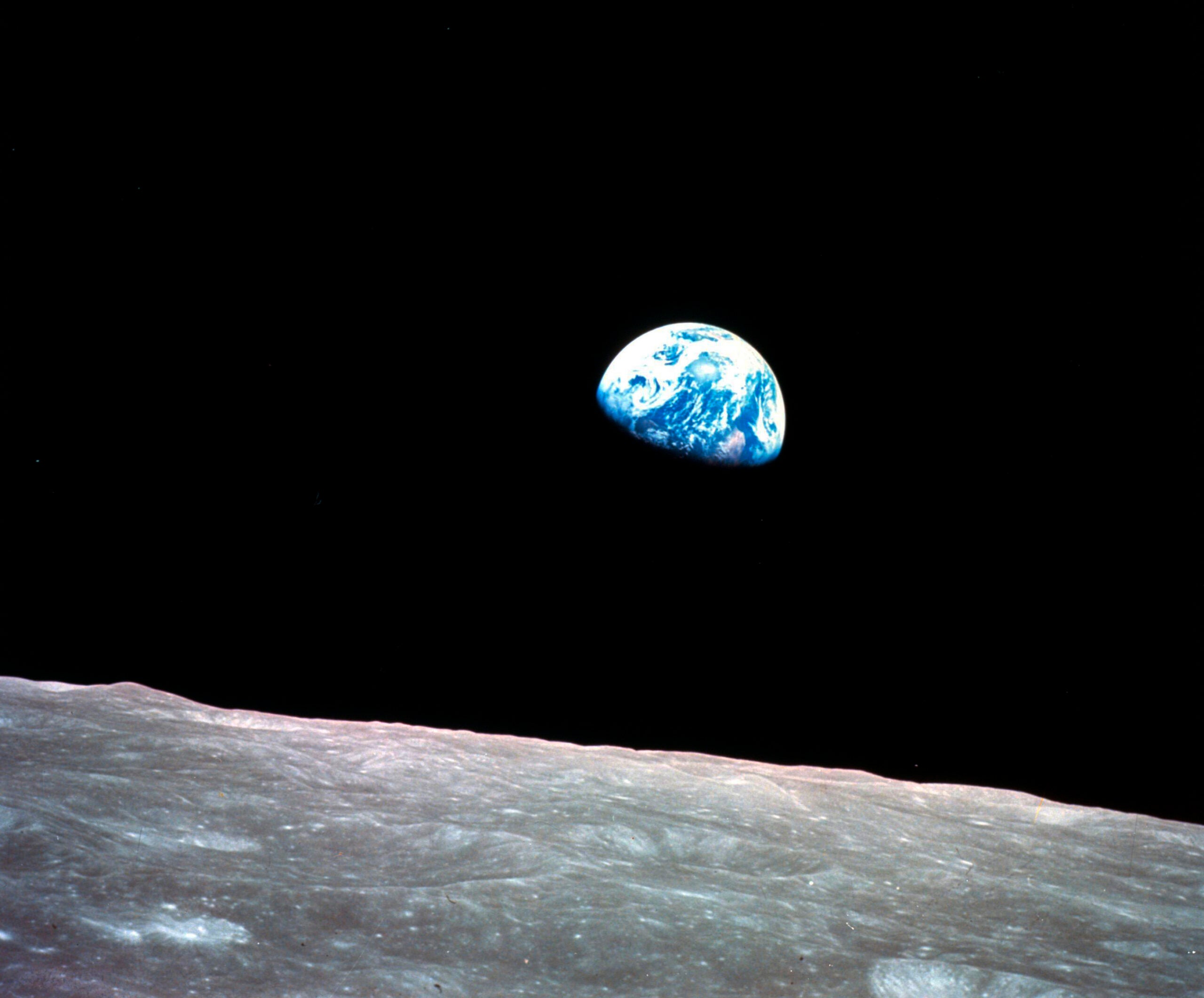 View of Earth rising over the lunar horizon, with the moons surface in the foreground and the planet partially visible against the blackness of space.