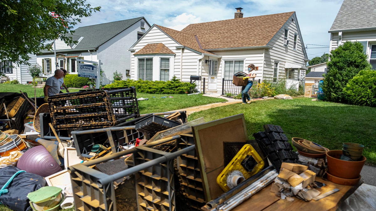 People remove damaged belongings from a house, placing items on the lawn and curb. The scene shows discarded household goods outside suburban homes on a sunny day.