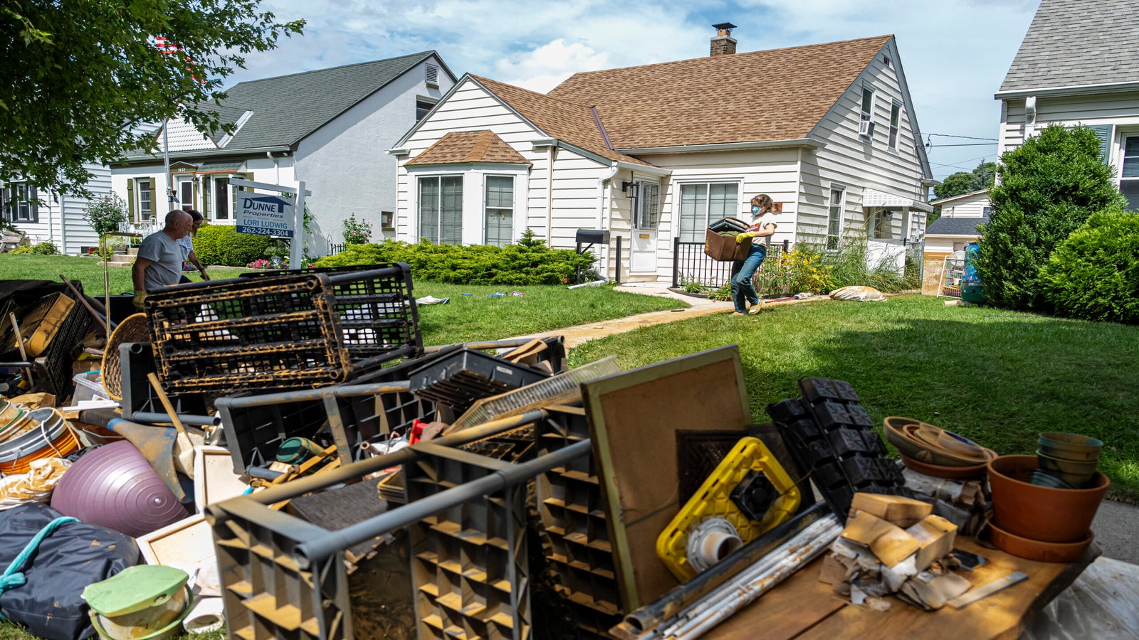 People remove damaged belongings from a house, placing items on the lawn and curb. The scene shows discarded household goods outside suburban homes on a sunny day.
