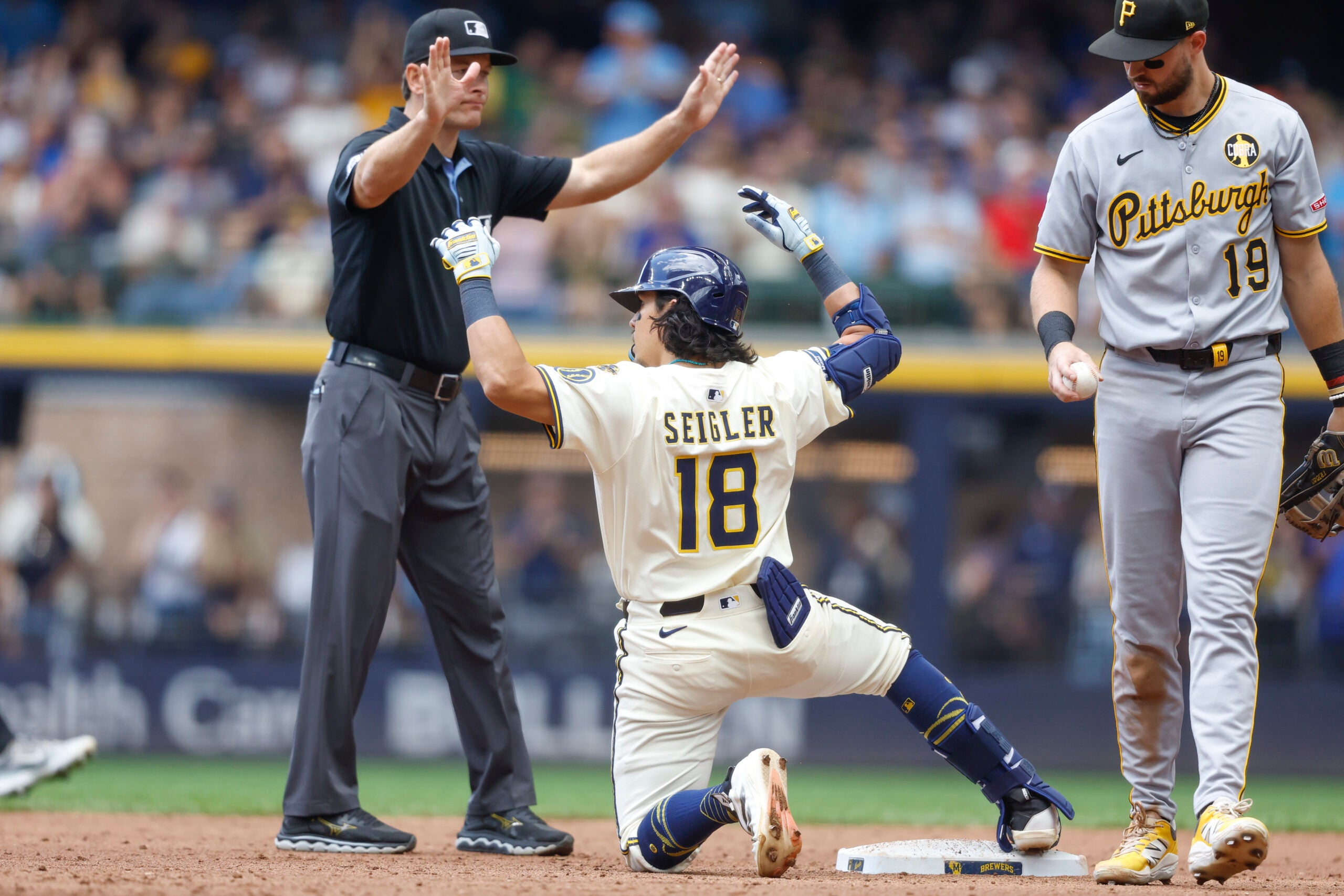 A baseball umpire signals safe as a player in a white uniform kneels at a base, while an opposing player in a gray uniform stands nearby.