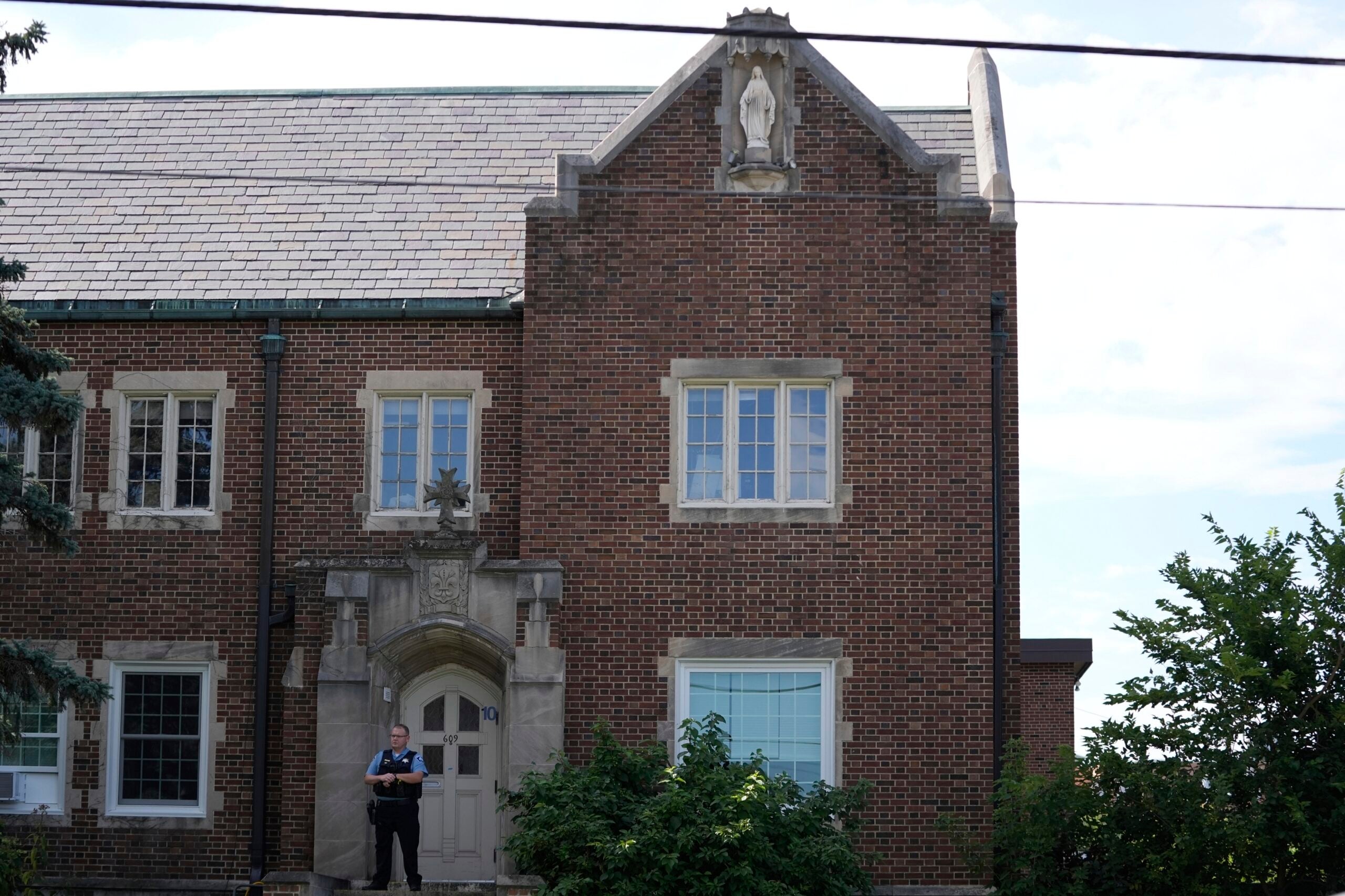 A police officer stands outside a brick building with stone-framed windows and a statue on the roof.