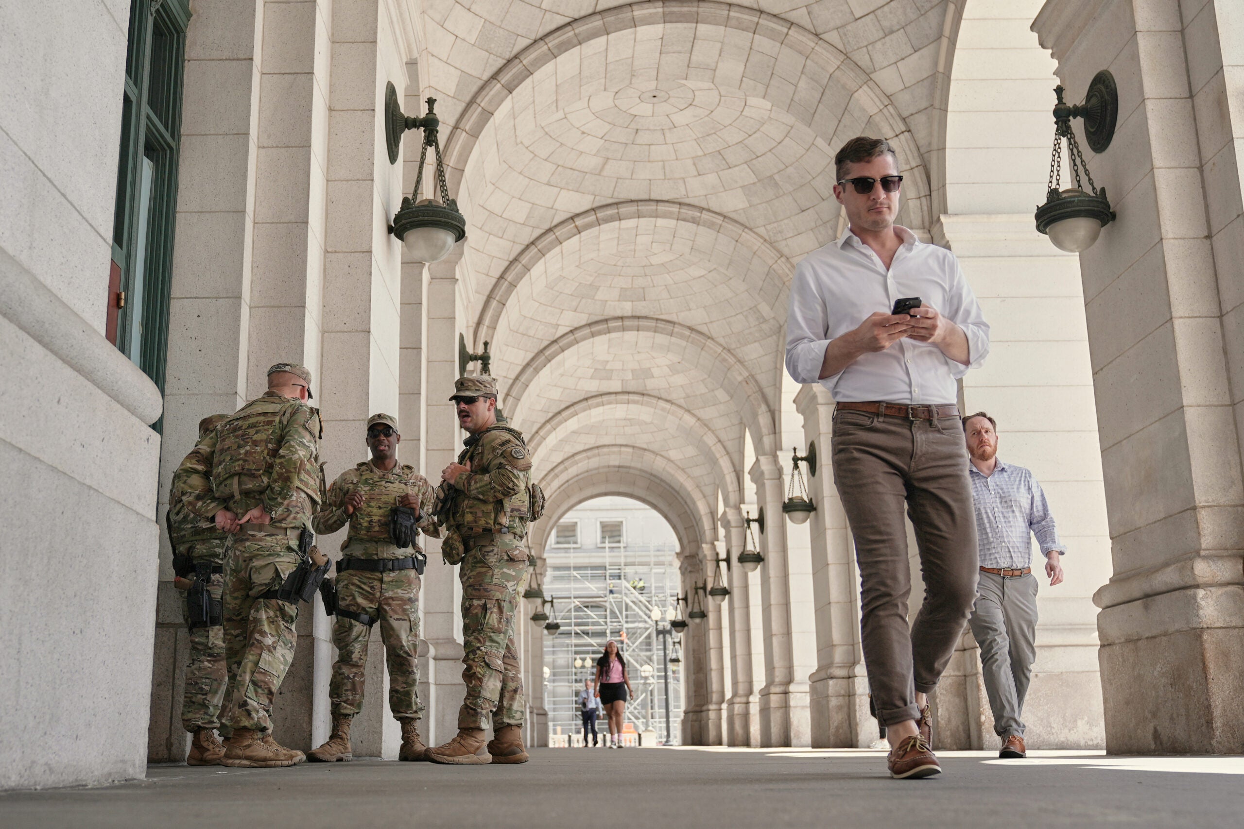 A group of soldiers stand talking under an arched walkway while a man in casual clothes walks by, looking at his phone; other people walk in the background.