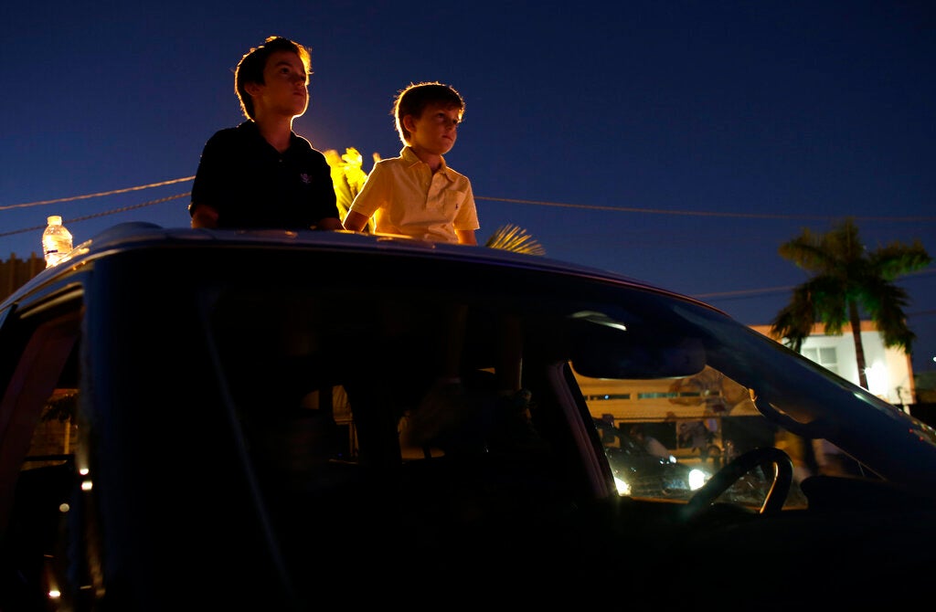 Two boys stand through the sunroof of a car at dusk, illuminated by a light source behind them, with buildings and palm trees visible in the background.