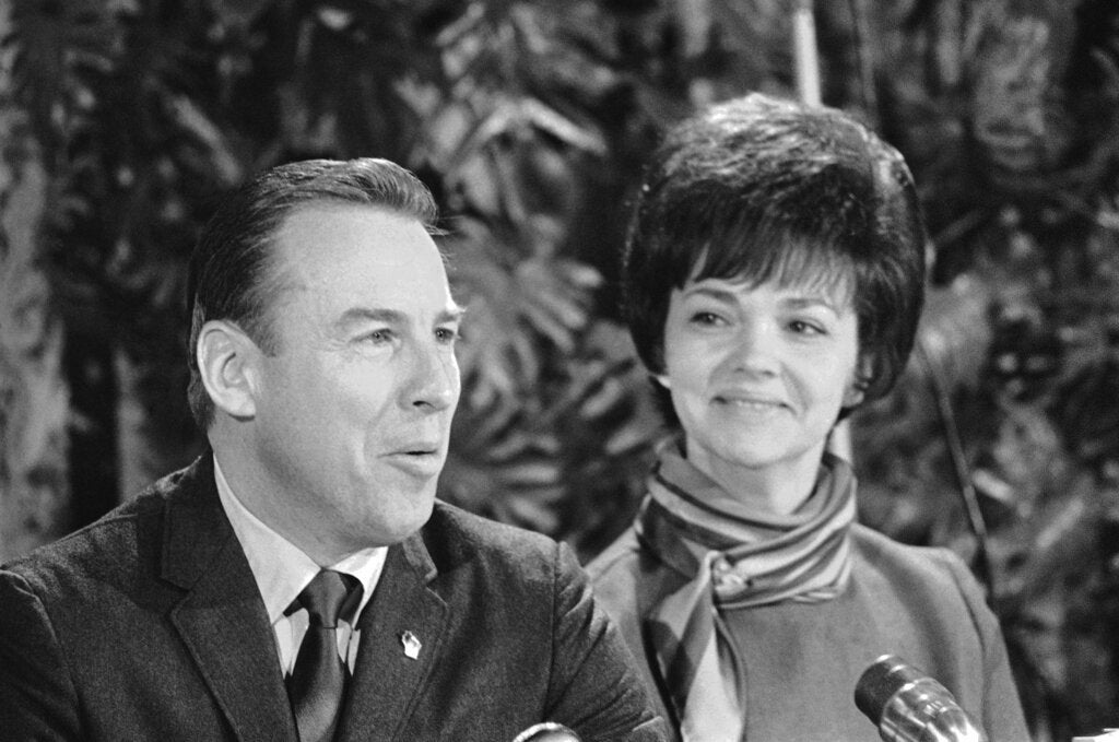 A man in a suit speaks into microphones while a woman beside him smiles, both seated indoors with foliage in the background.