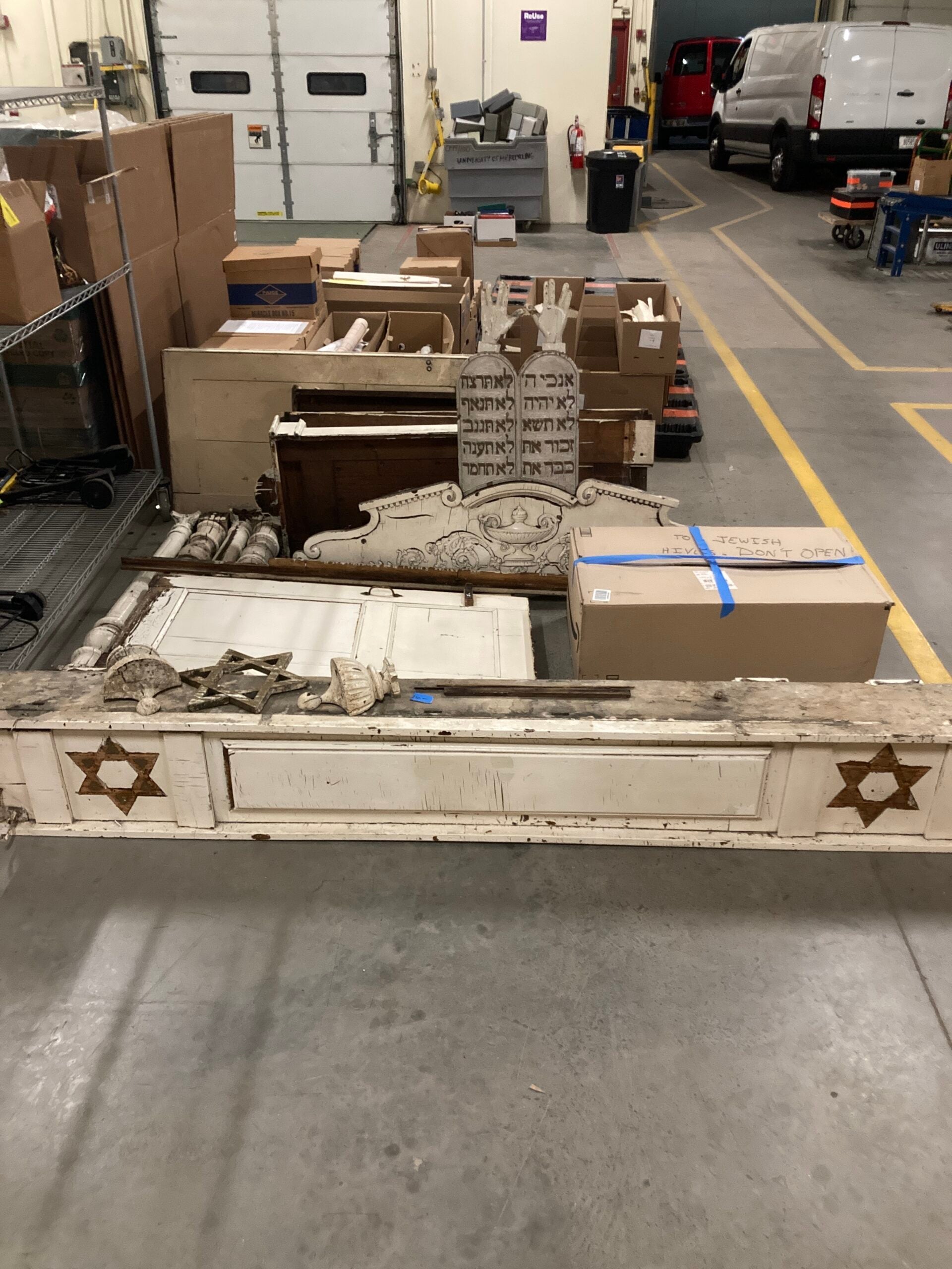 Several dismantled wooden artifacts with Hebrew inscriptions and Star of David symbols are laid out on the floor in a warehouse, surrounded by packed boxes and vehicles.