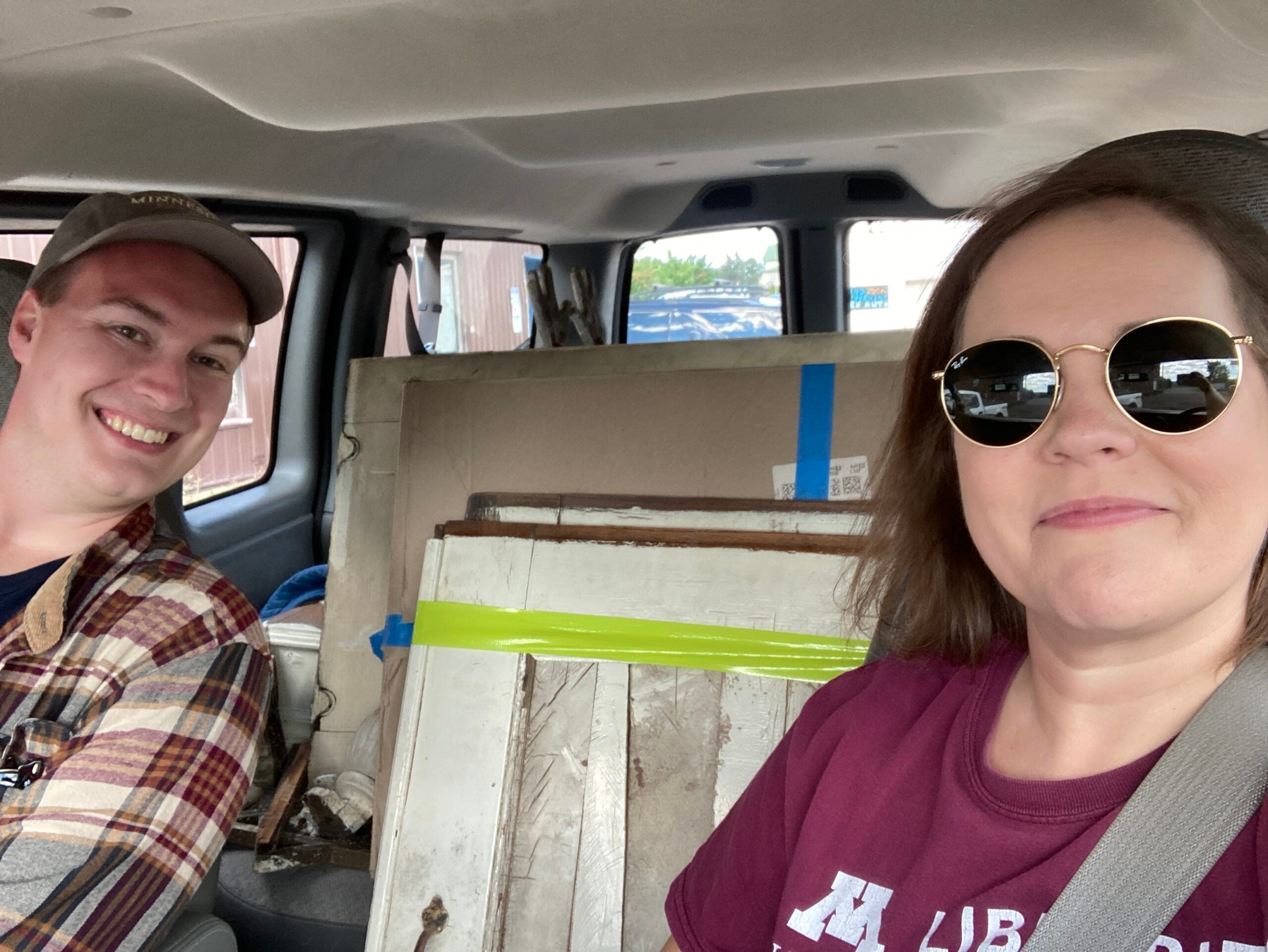 Two people sit in a vehicle with pieces of wood and other materials secured behind them. Both are smiling; the woman wears sunglasses and a maroon shirt, the man wears a plaid shirt and cap.