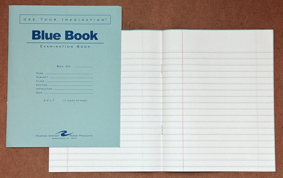A blue examination book labeled Blue Book is shown closed next to an open lined notebook on a brown surface.