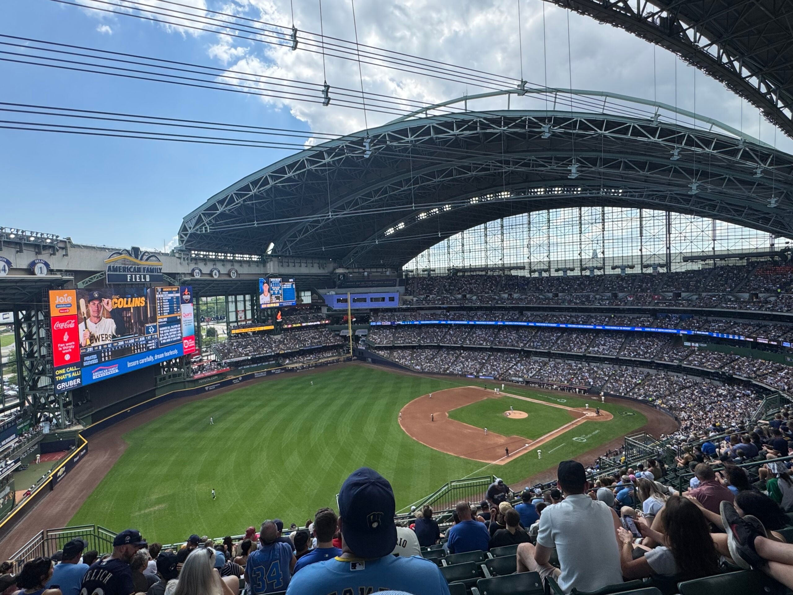 Wide-angle view of a baseball game at an indoor stadium with a partially open roof, large crowd in attendance, and scoreboard visible on the left.