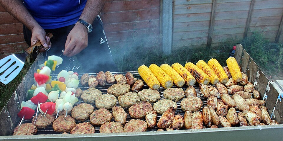 A person grills assorted kebabs, chicken patties, wings, and corn on the cob on a barbecue grill outdoors.
