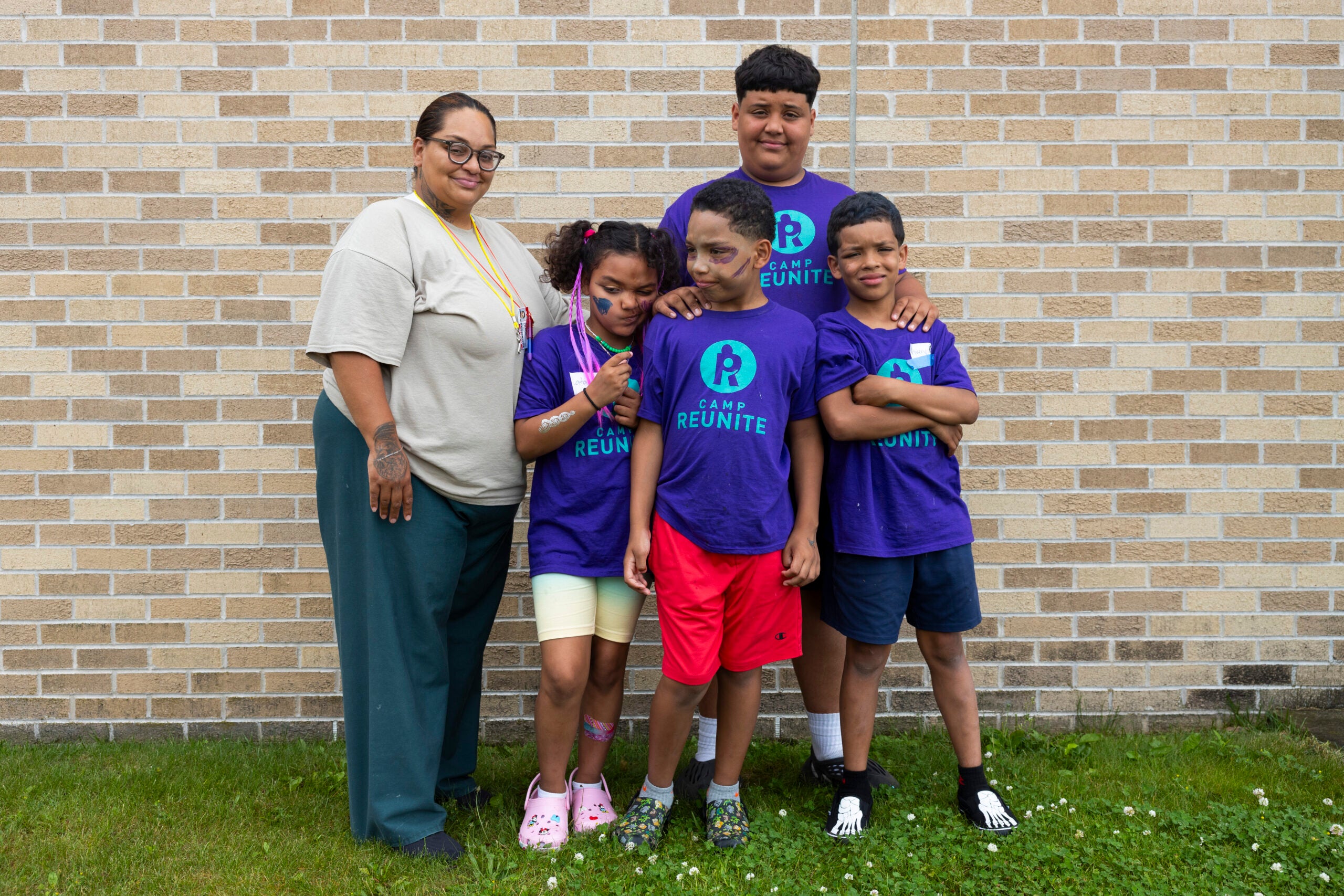 A woman stands with four children wearing matching purple Camp Reunite shirts, posing in front of a tan brick wall on green grass.