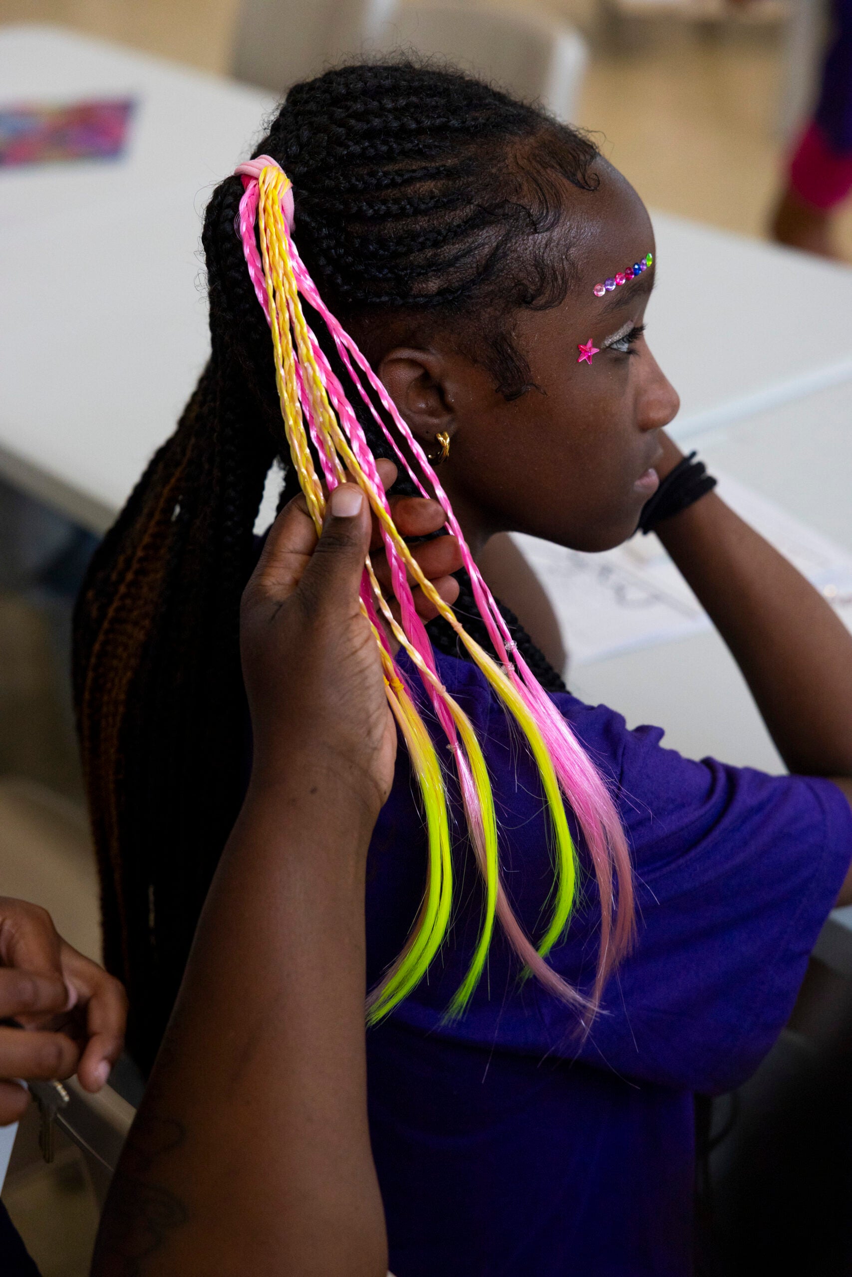 A person holds the braided hair of a girl with pink and yellow extensions and decorative face gems as she sits at a table.