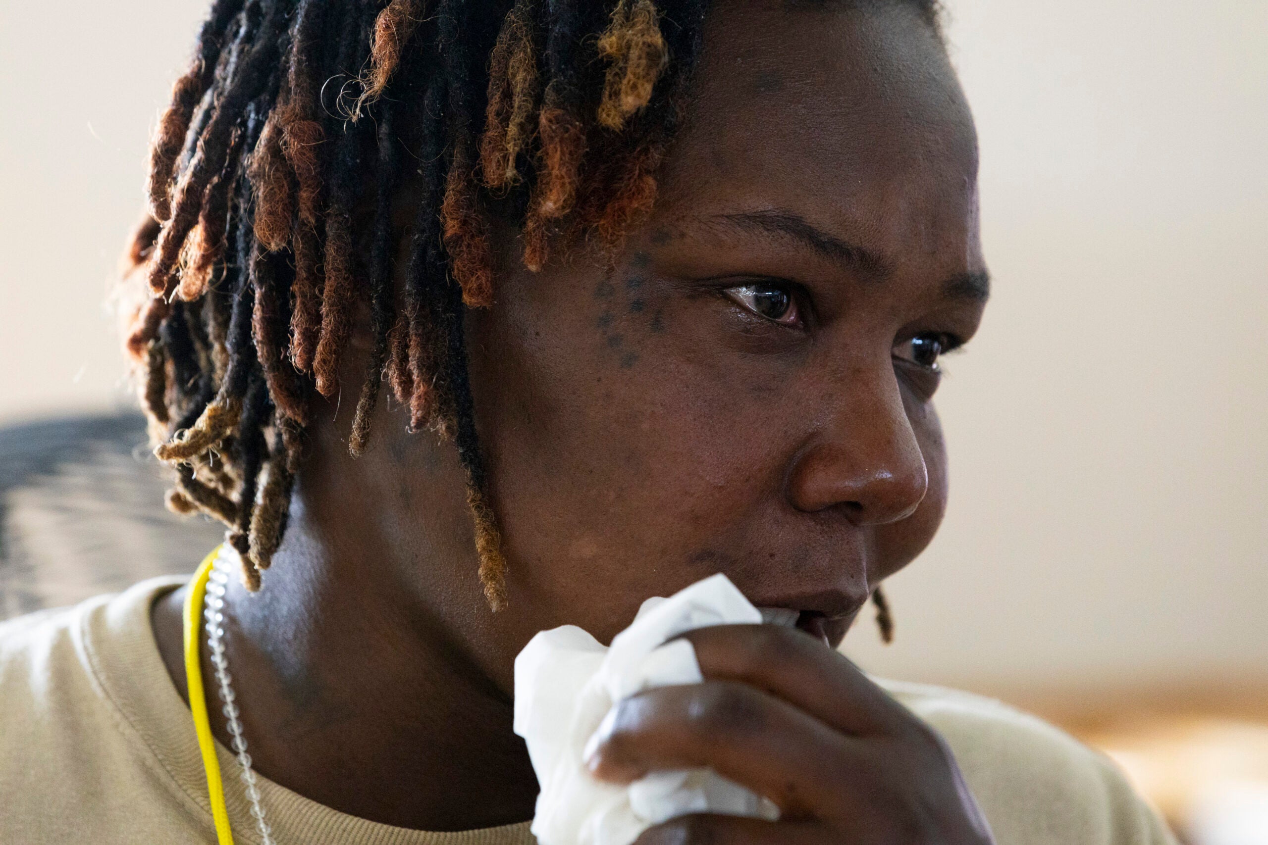 A person with short, dreadlocked hair holds a tissue to their mouth, appearing emotional with tears in their eyes.