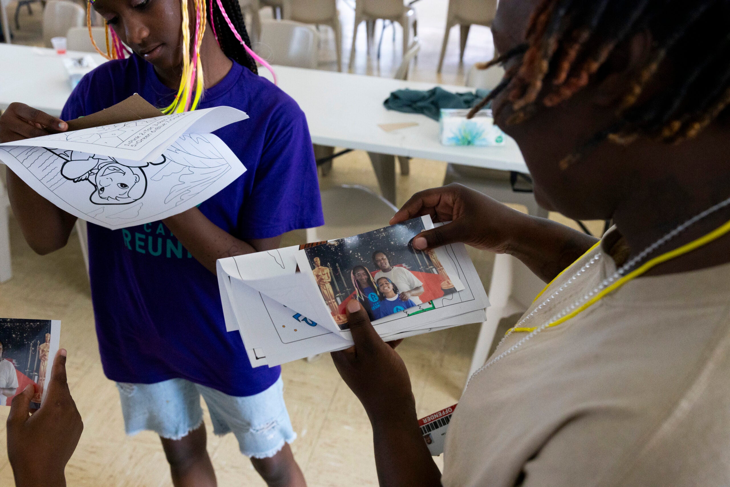 Two people look at printed photos and coloring pages while standing near a table in a room with empty chairs.