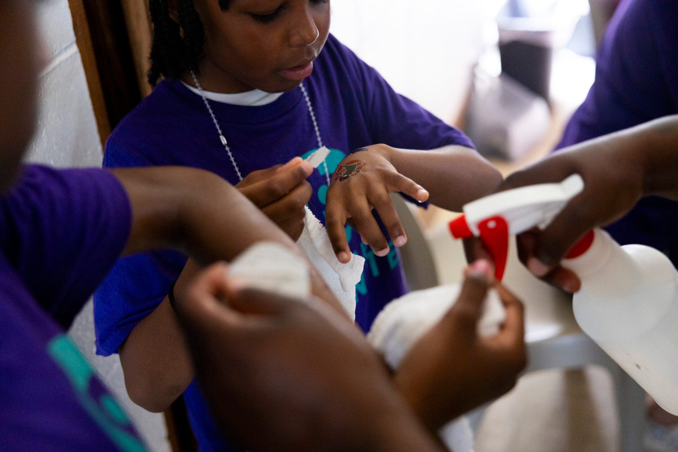 A child applies a bandage to their hand while others assist with a spray bottle and cloth.