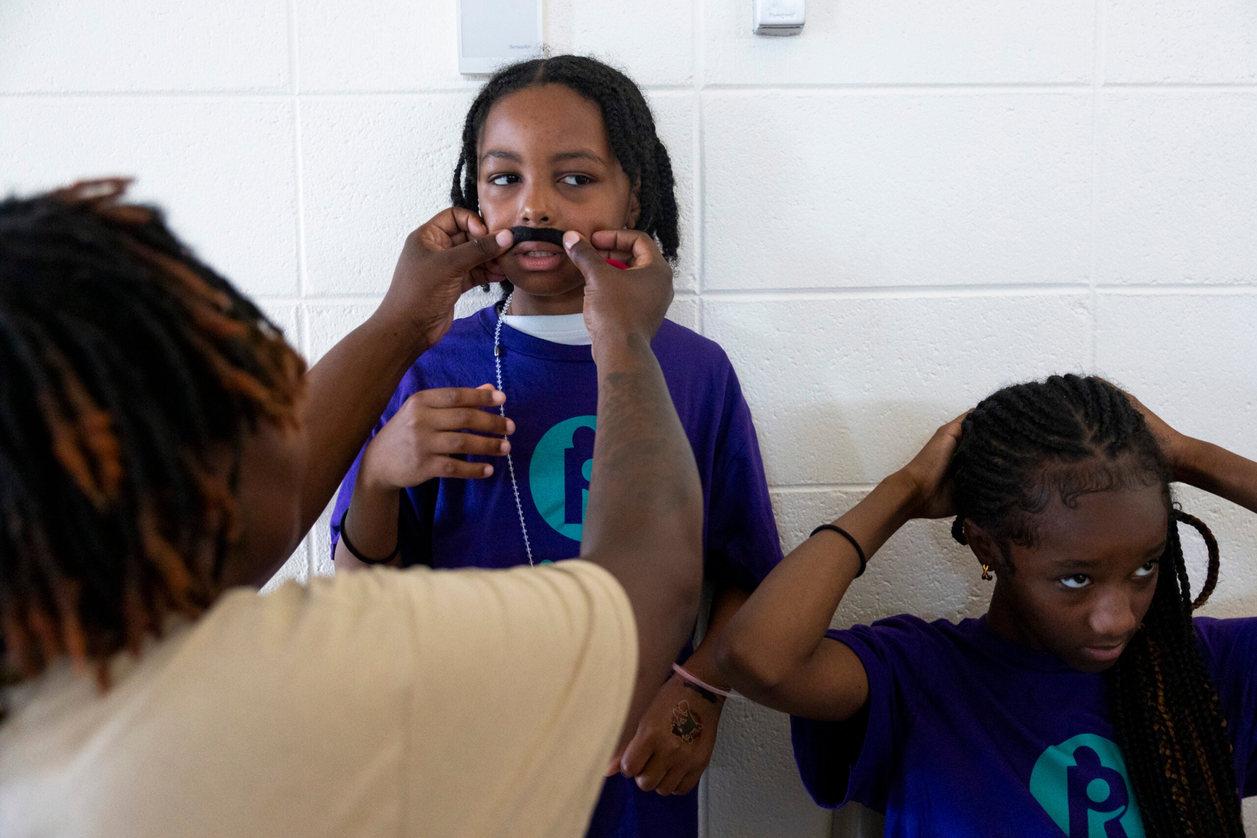 A person adjusts a childs fake mustache while another child fixes her hair; both children wear matching purple shirts and stand against a white wall.