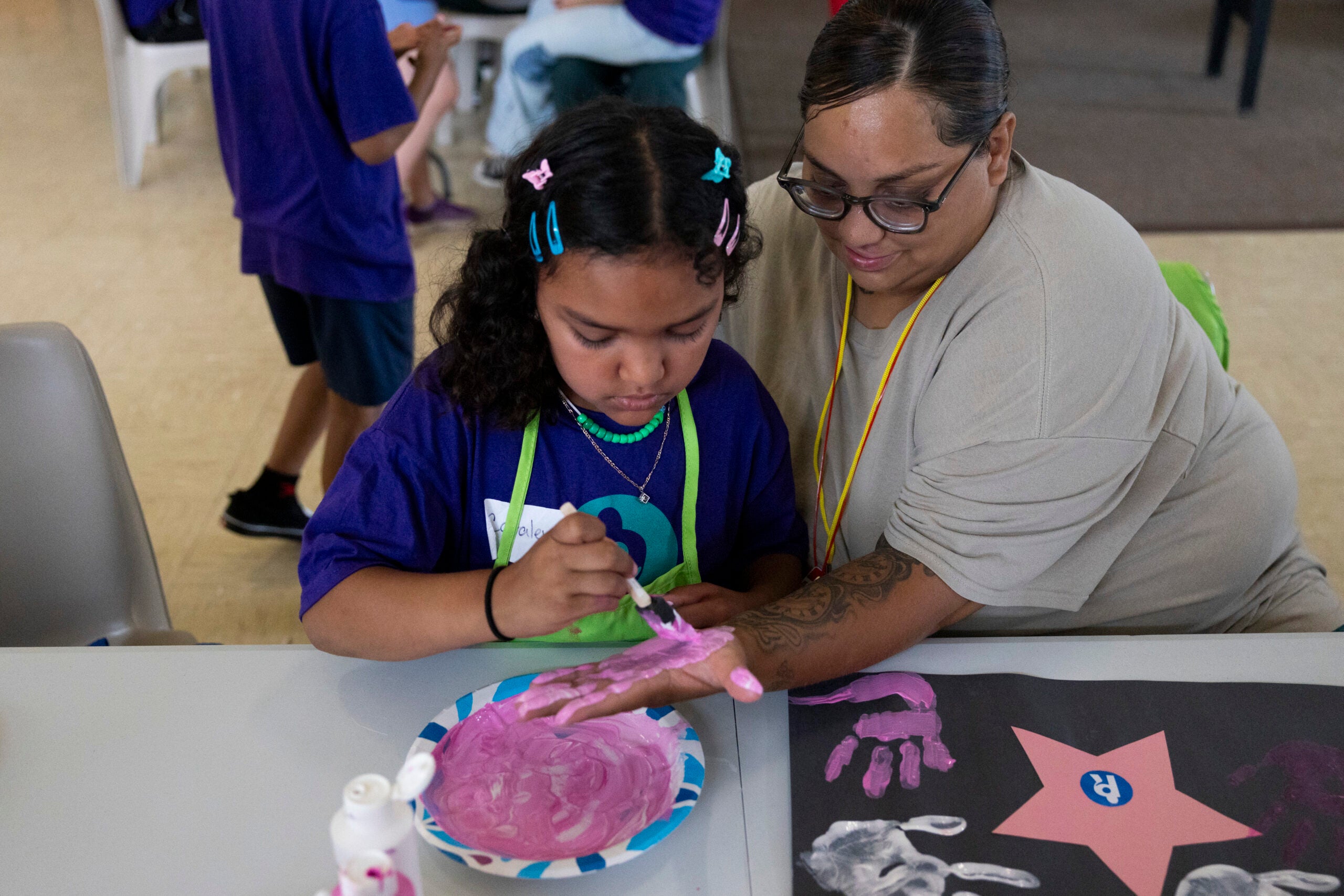 An adult helps a child paint their hand with pink paint at a table, preparing to make a handprint on black paper.