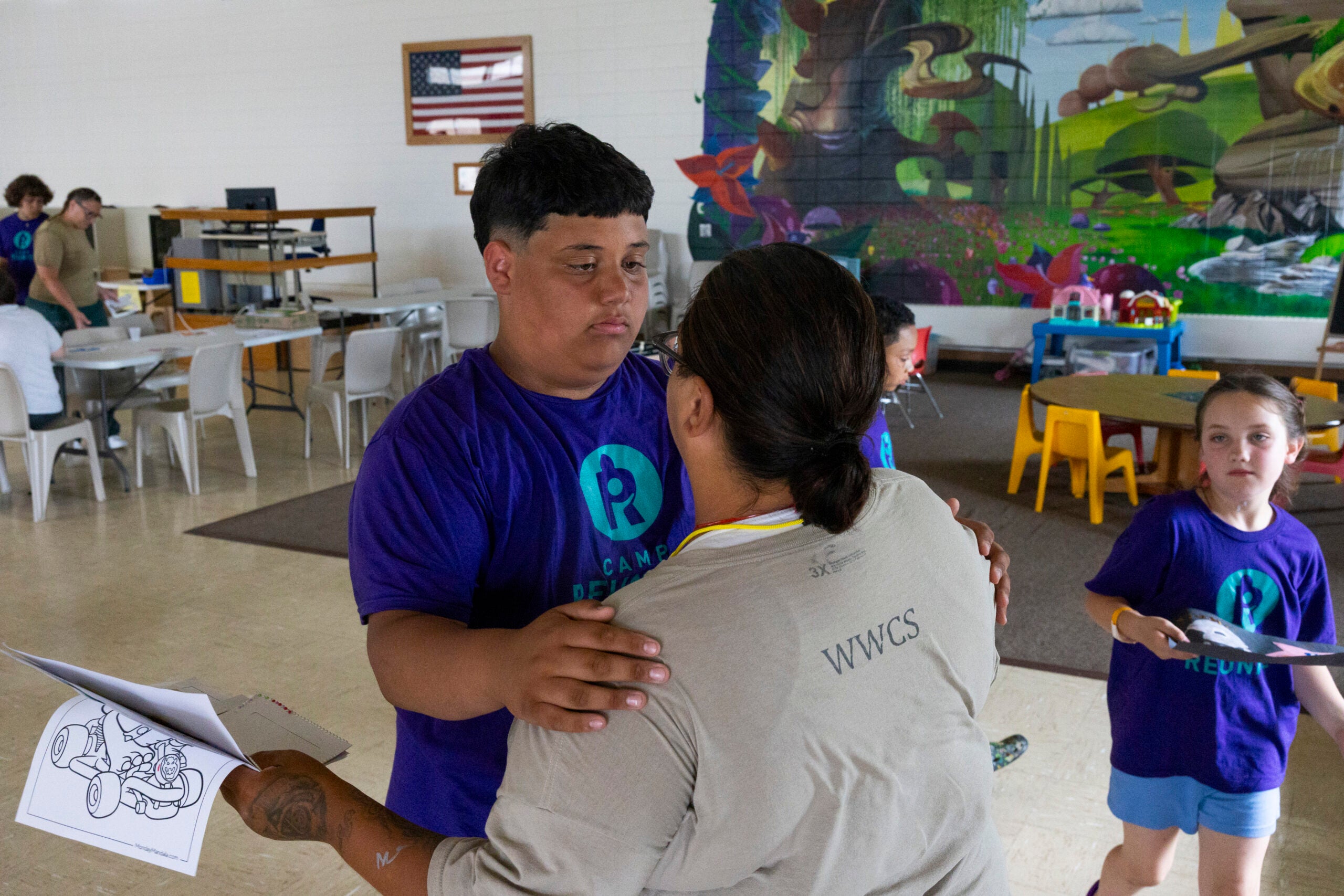 A staff member embraces a boy holding a coloring page in a classroom with other children and a mural on the wall.