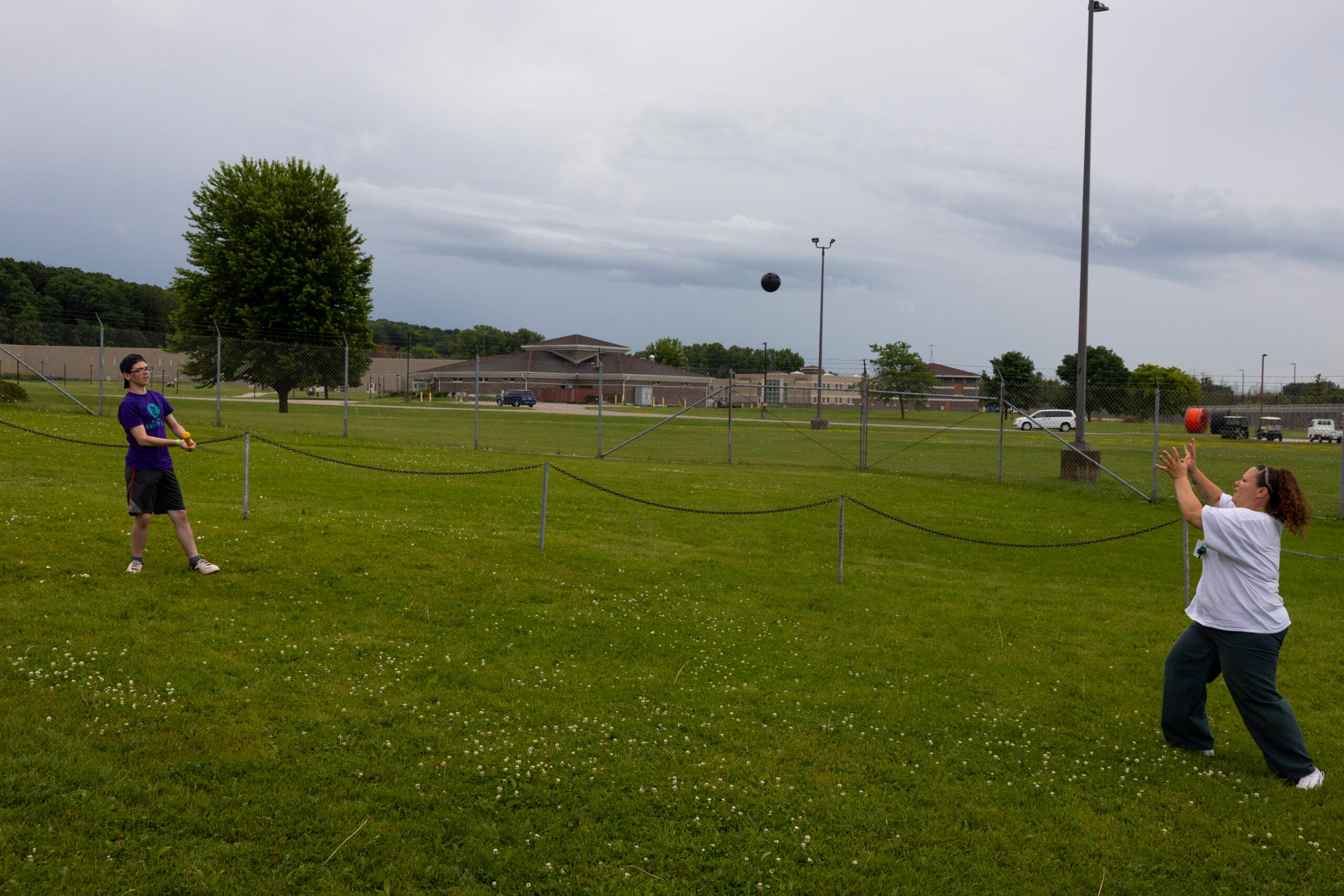 Two people play tetherball on a grassy field, one striking the ball and the other preparing to catch it; buildings and parked cars are visible in the background.