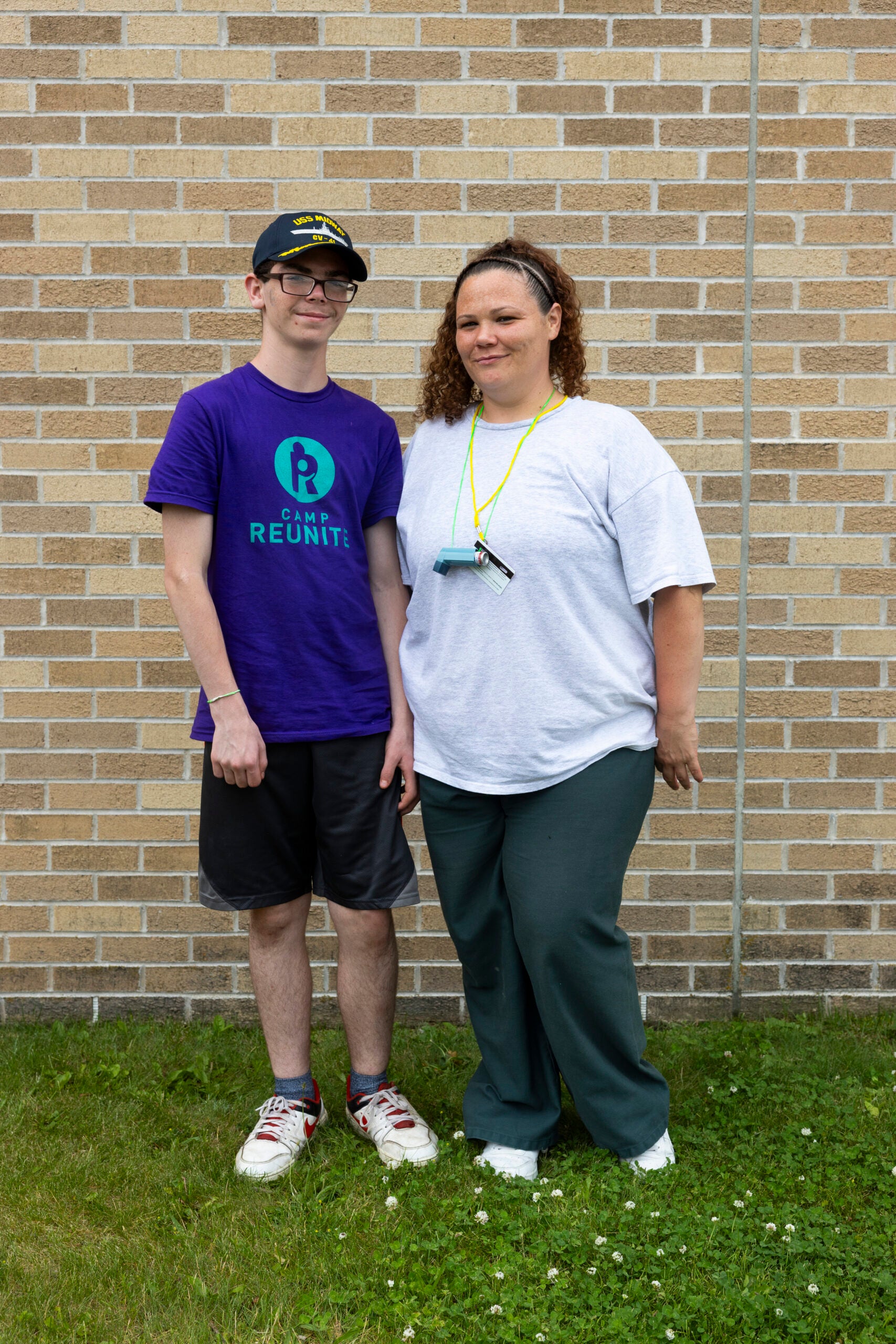 A woman and a teenage boy stand side by side on grass in front of a brick wall, posing for a photo.