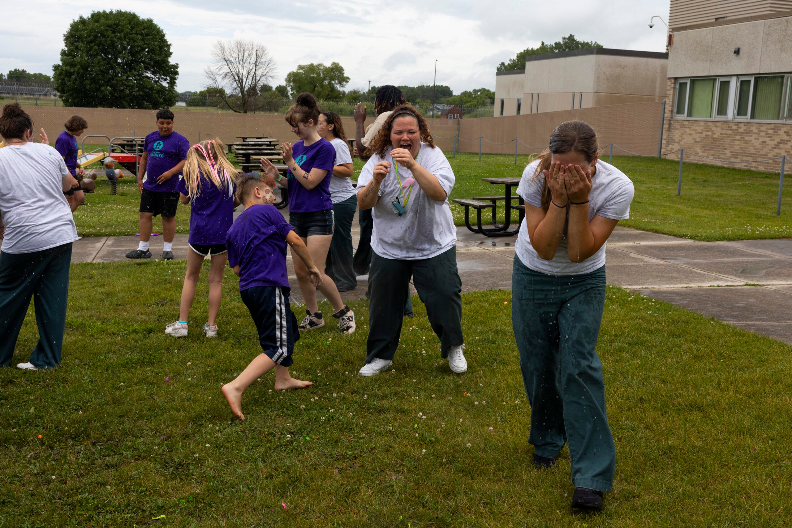 A group of people, including children and adults, participate in an outdoor activity on grass near a building; some are laughing and covering their faces while water is being sprayed.