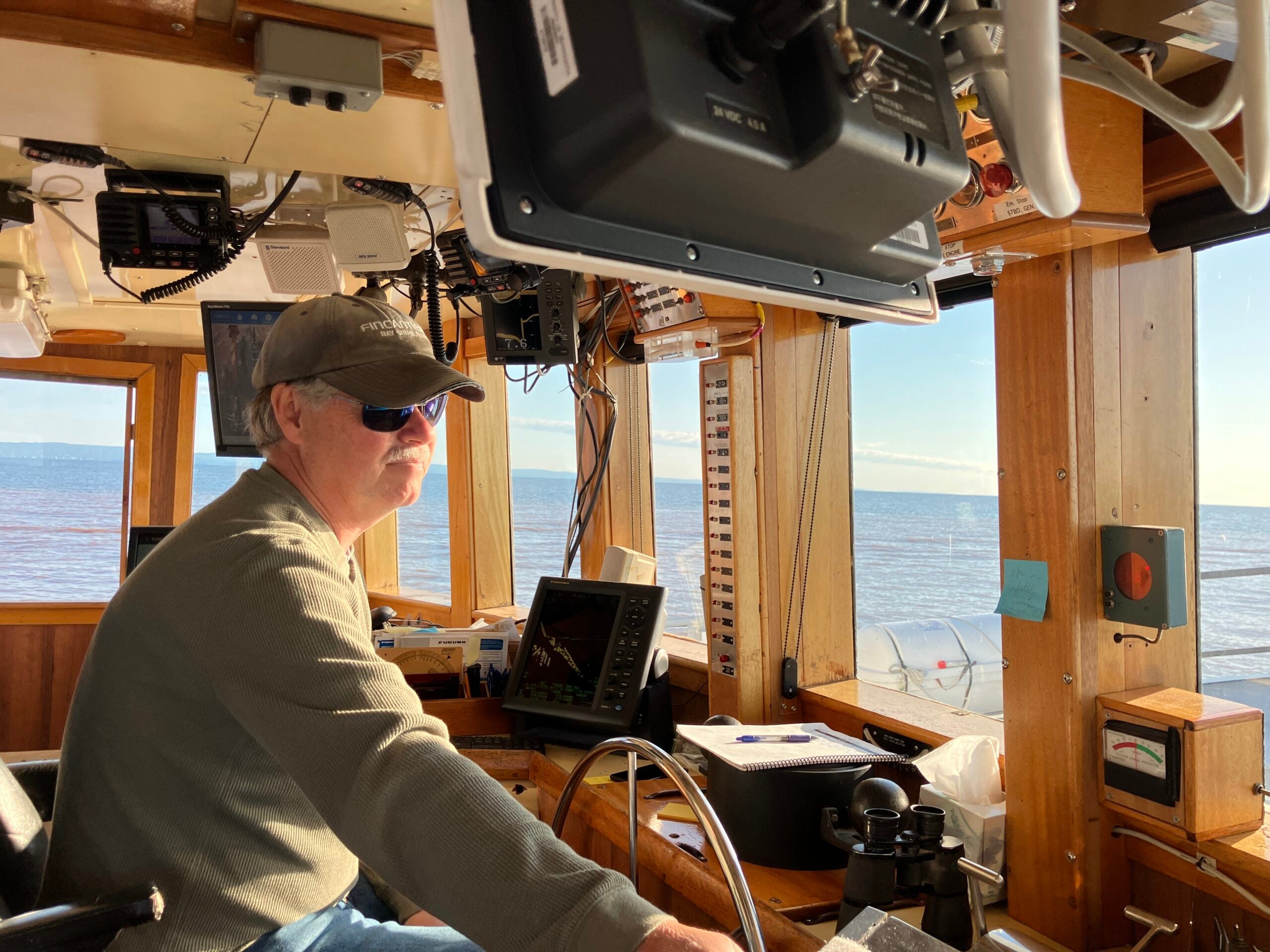 Man wearing sunglasses and a cap operates the wheel of a boat in a sunlit wooden cabin, with navigational equipment and an ocean view visible through the windows.