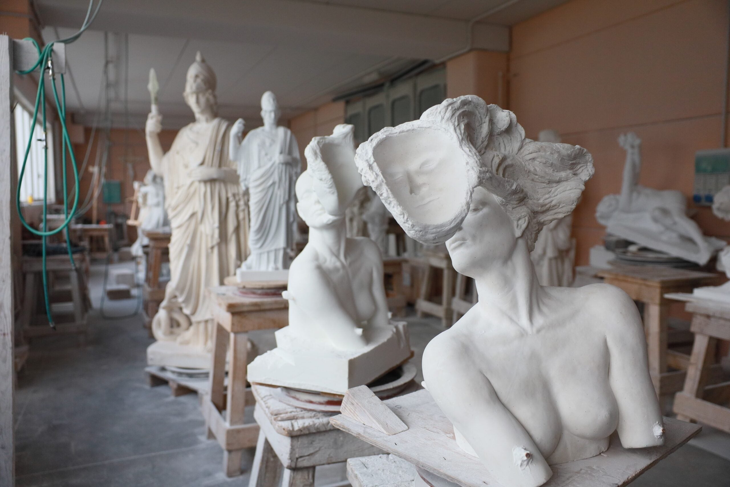 Several unfinished plaster sculptures, including fragmented busts and full statues, are displayed on wooden work tables in an art studio.