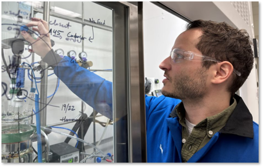 A man wearing safety glasses and a blue lab coat adjusts equipment inside a laboratory fume hood with handwritten notes on the glass.