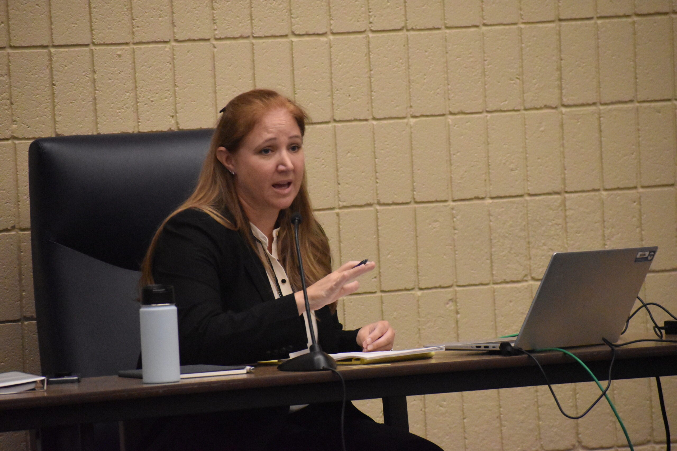 A woman in professional attire speaks at a desk with a laptop, water bottle, papers, and microphone in front of her against a beige brick wall.