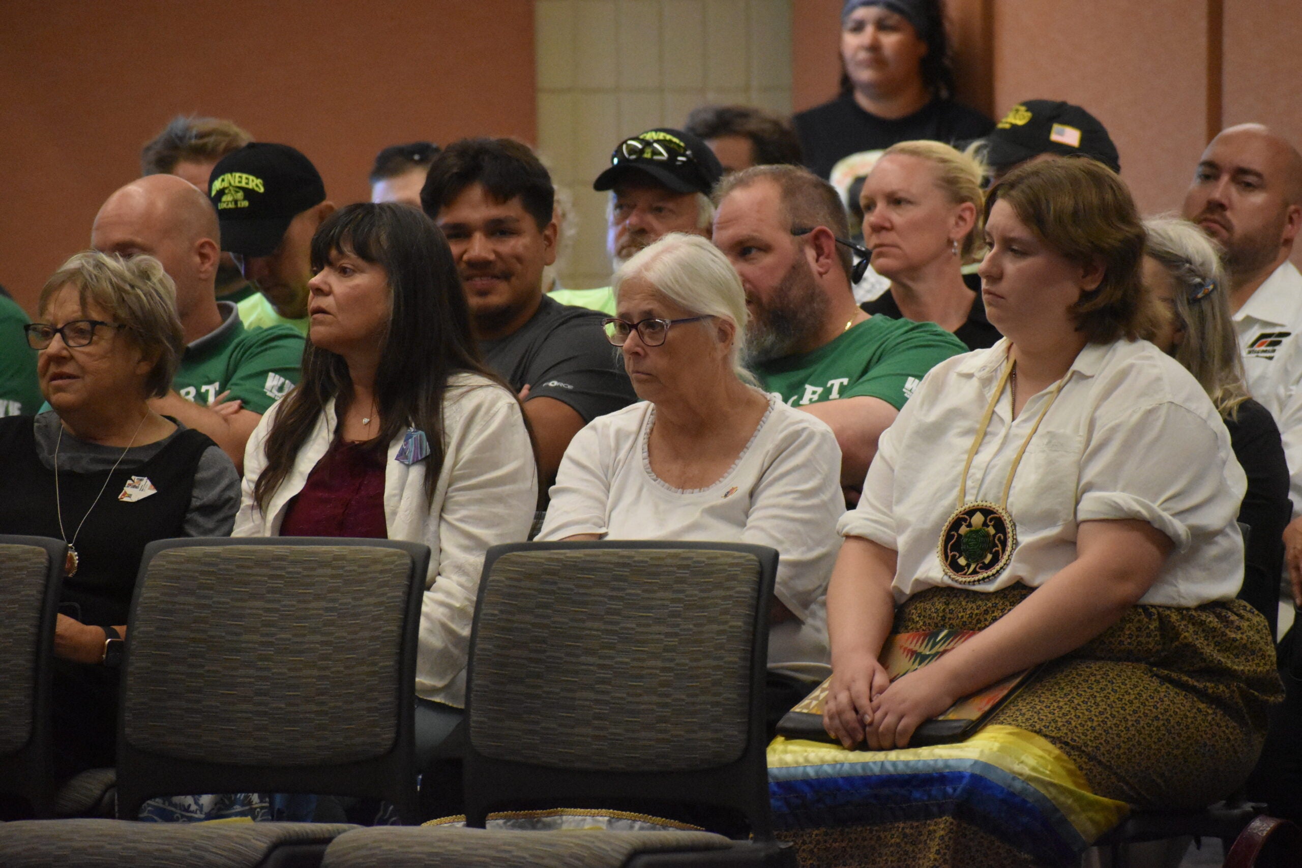 A group of people sit in rows of chairs at an indoor event, listening attentively. Some attendees wear casual clothes, while others wear traditional attire.