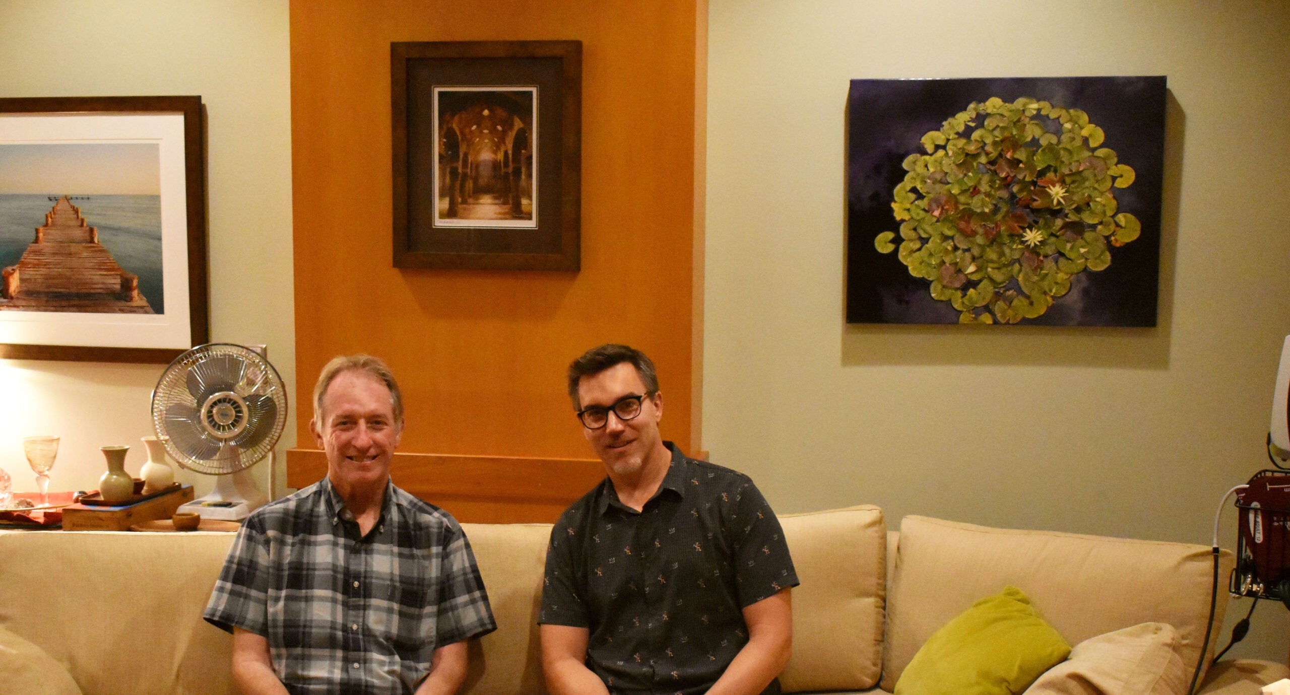 Two men are sitting on a beige couch in a living room with framed artwork and a fan on the wall behind them.