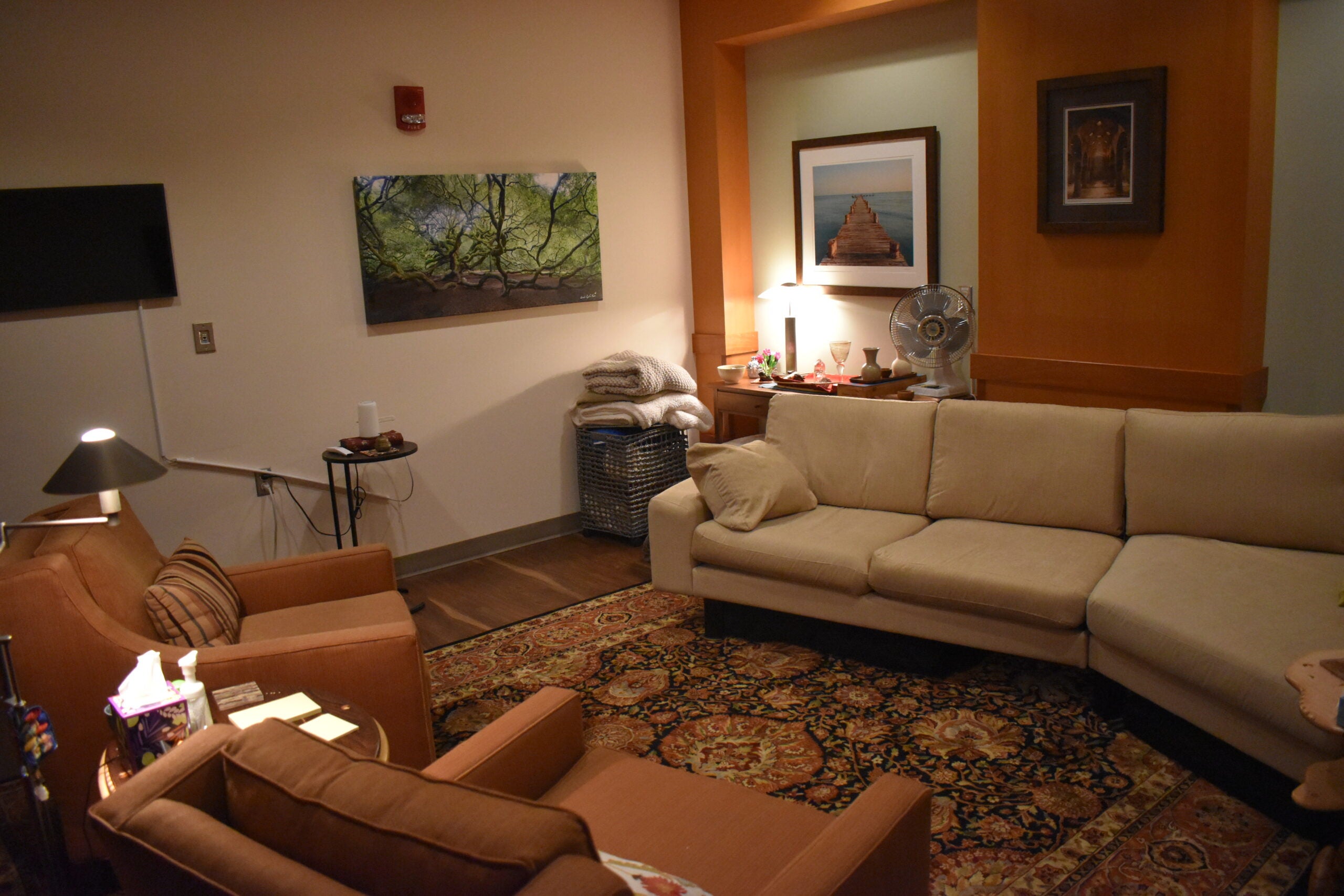 A cozy living room with beige sofas, an armchair, a patterned rug, wall art, a lamp, and folded blankets on a shelf in the corner.