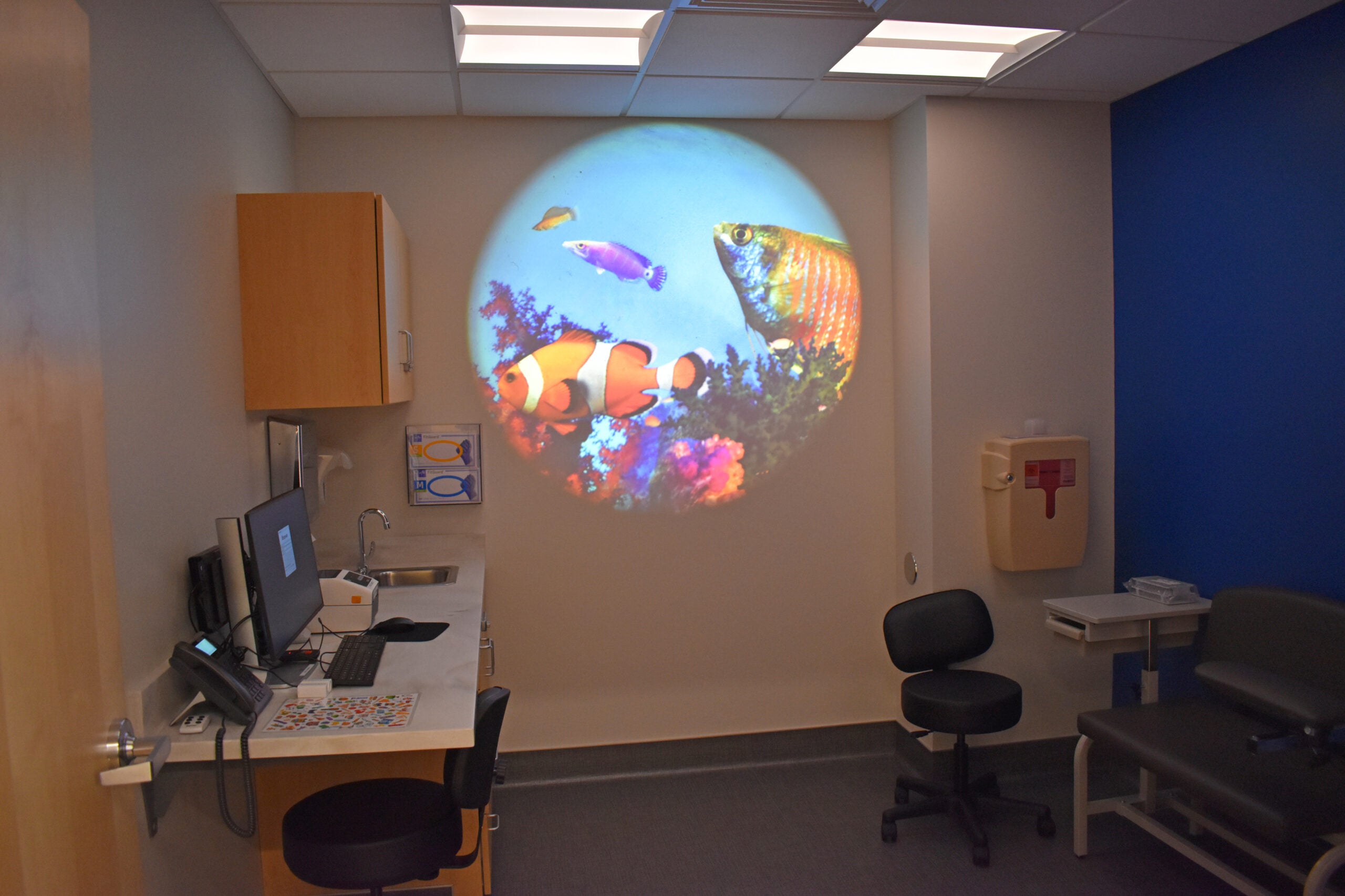 A doctor’s office exam room with a computer and medical equipment, featuring a round projection of colorful fish on the wall, and blue accent walls.