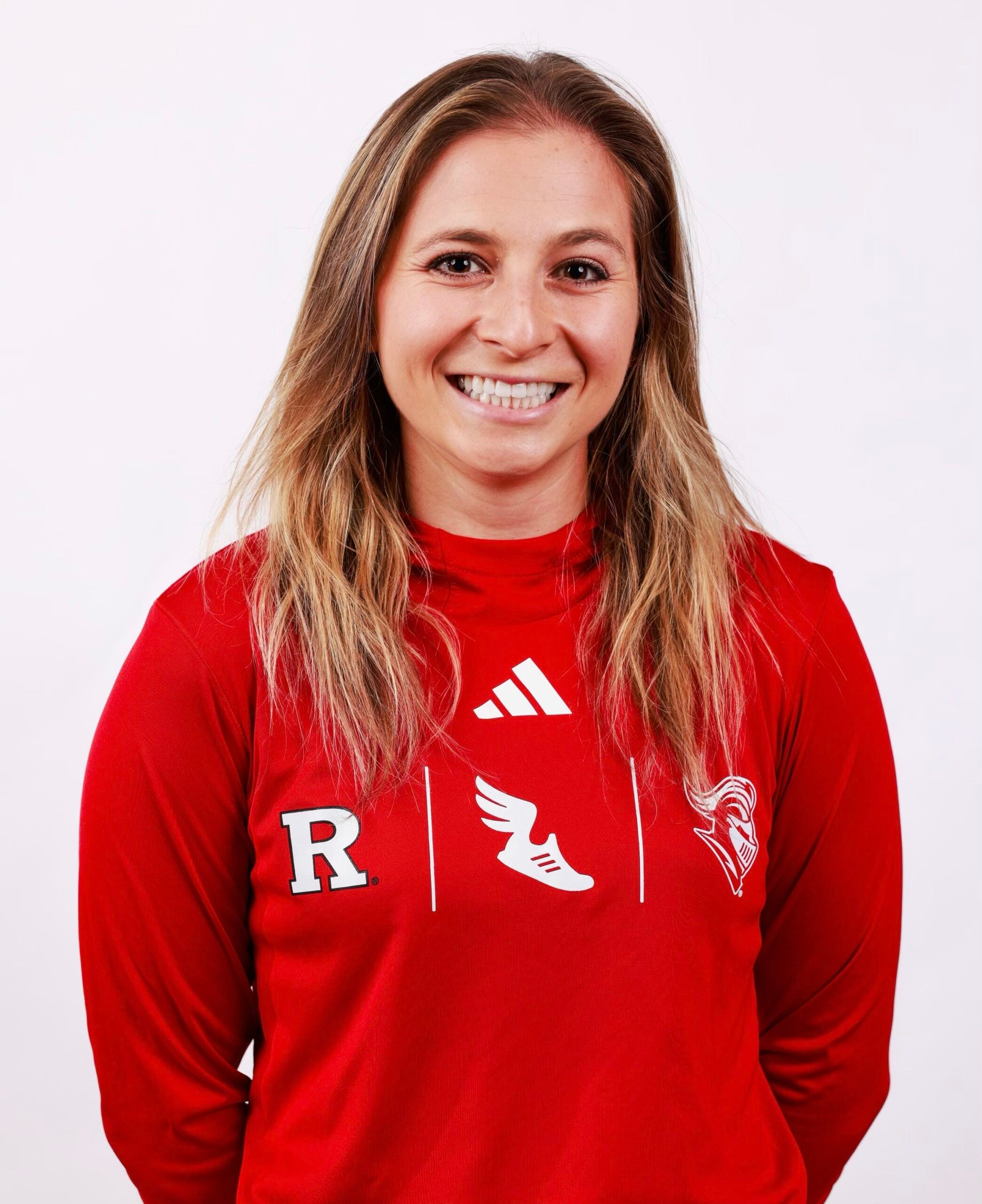 A woman with long blond hair smiles, wearing a red sports jersey with white logos and a letter R on the front, standing against a plain white background.